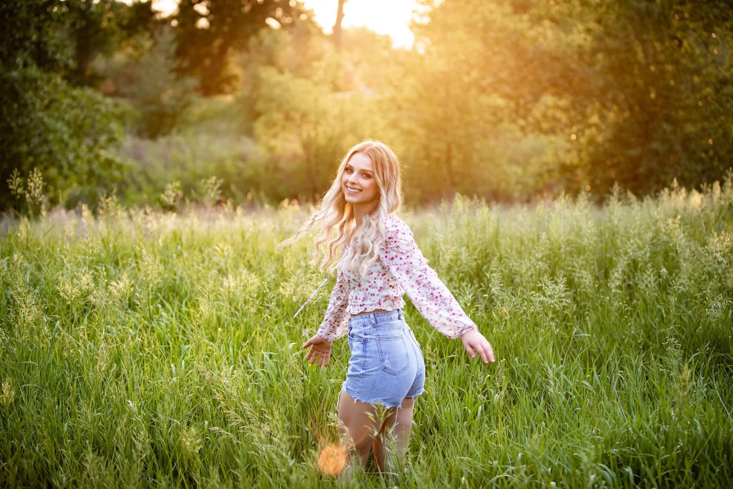 A young woman with long blonde curly hair smiling in a grassy field during sunset, wearing a floral blouse and denim shorts.