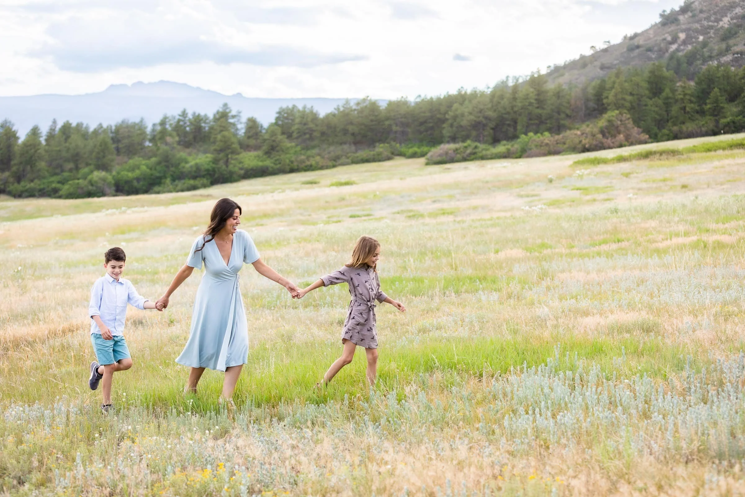 A woman and two children holding hands and walking through a grassy field with mountains and trees in the background.