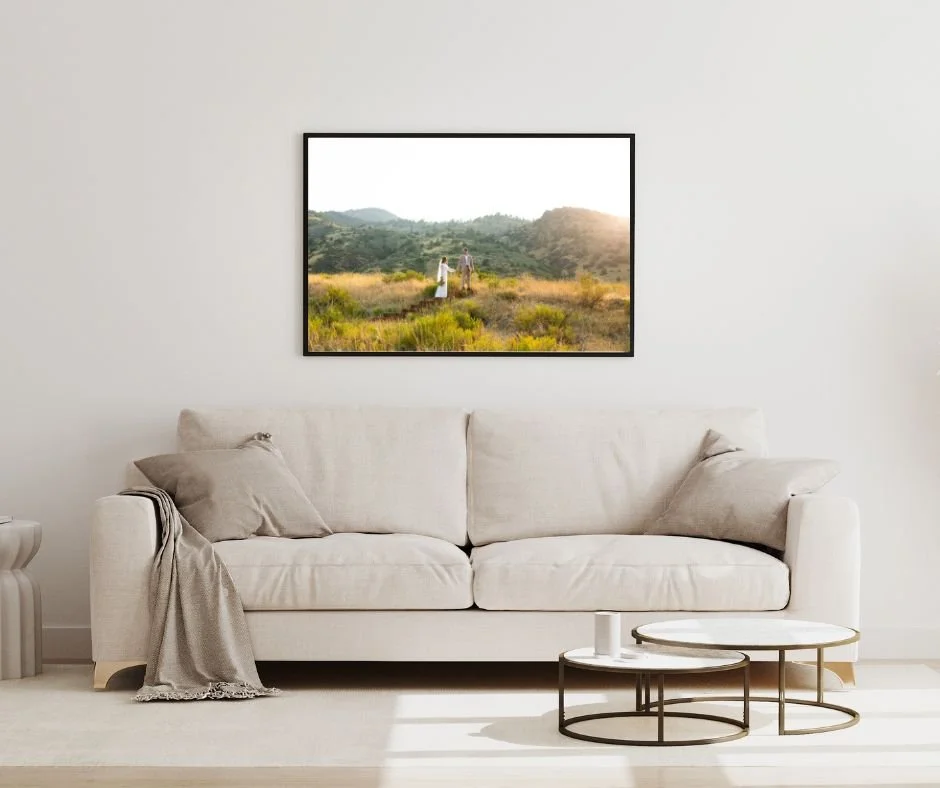 Living room with a beige sofa, pillows, a blanket, a round side table, and landscape painting of two people walking in a field at sunset.
