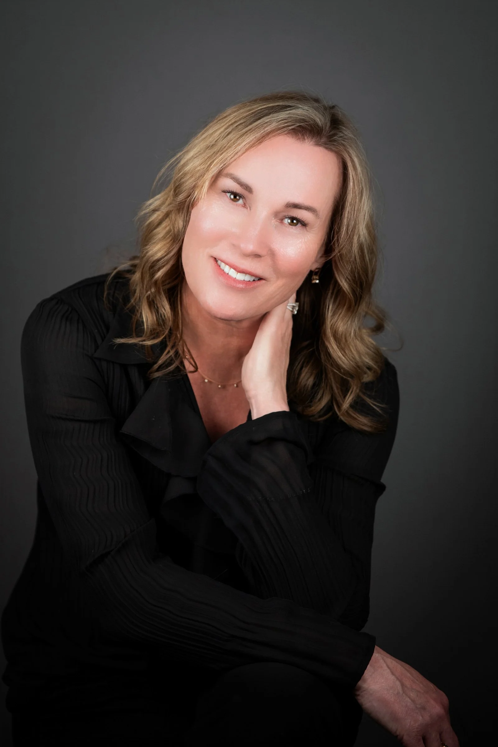 A woman with shoulder-length wavy blonde hair, wearing a black top, smiling against a dark gray background.