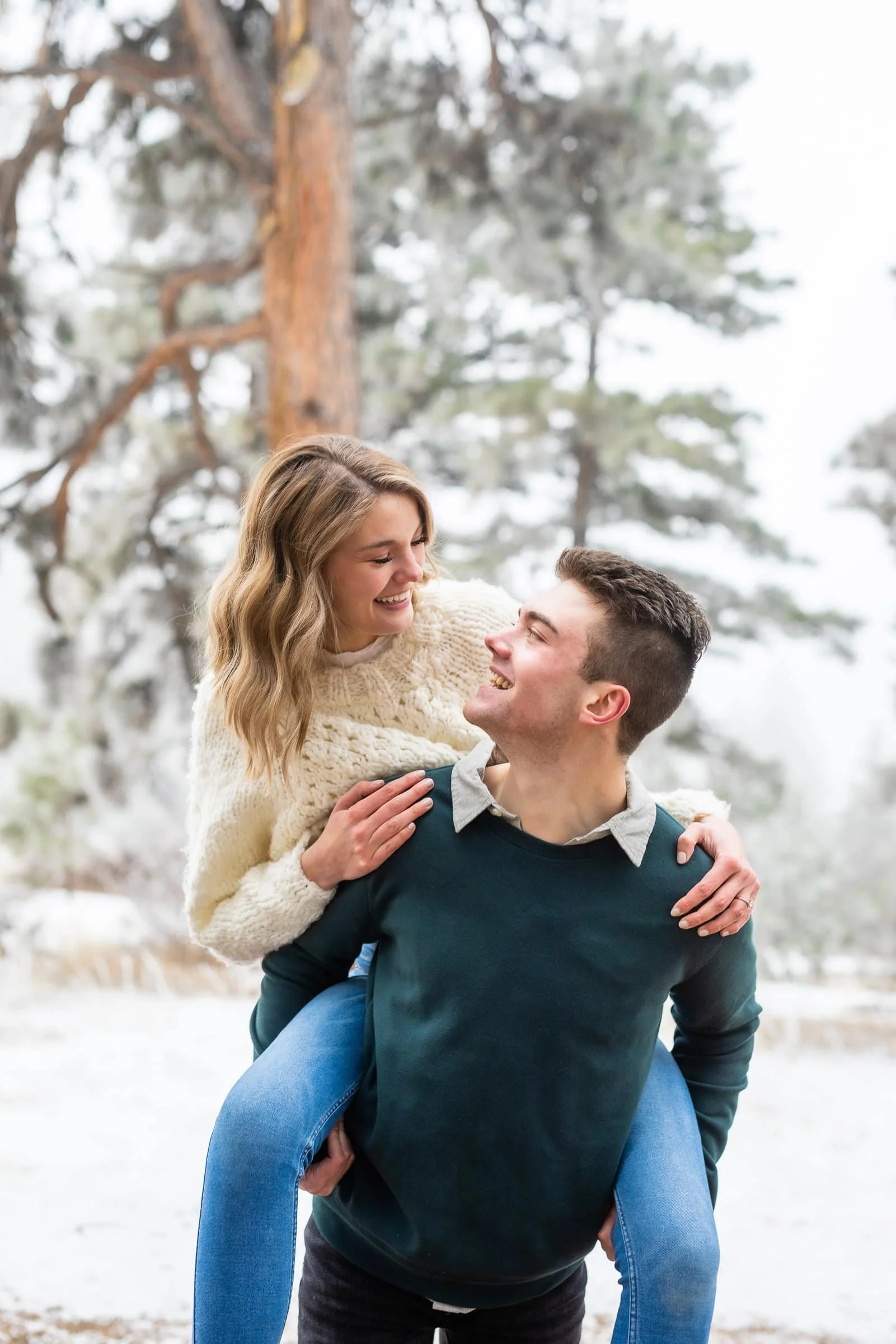 A happy couple outdoors in a snowy forest, with the man giving the woman a piggyback ride, both smiling and looking at each other.