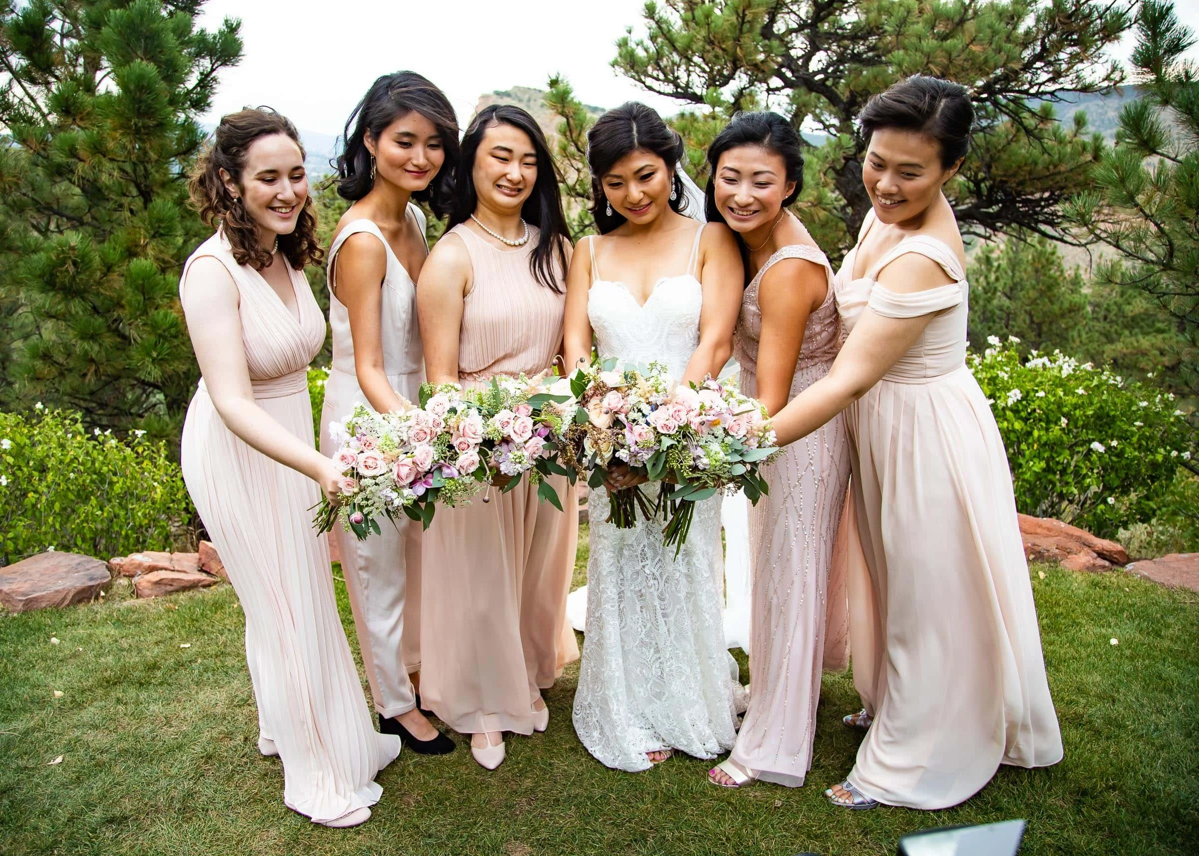 A bride in a white wedding dress with symbol lace stands with six bridesmaids in pastel-colored dresses outdoors, holding bouquets of pink and white flowers, surrounded by green trees and grass.