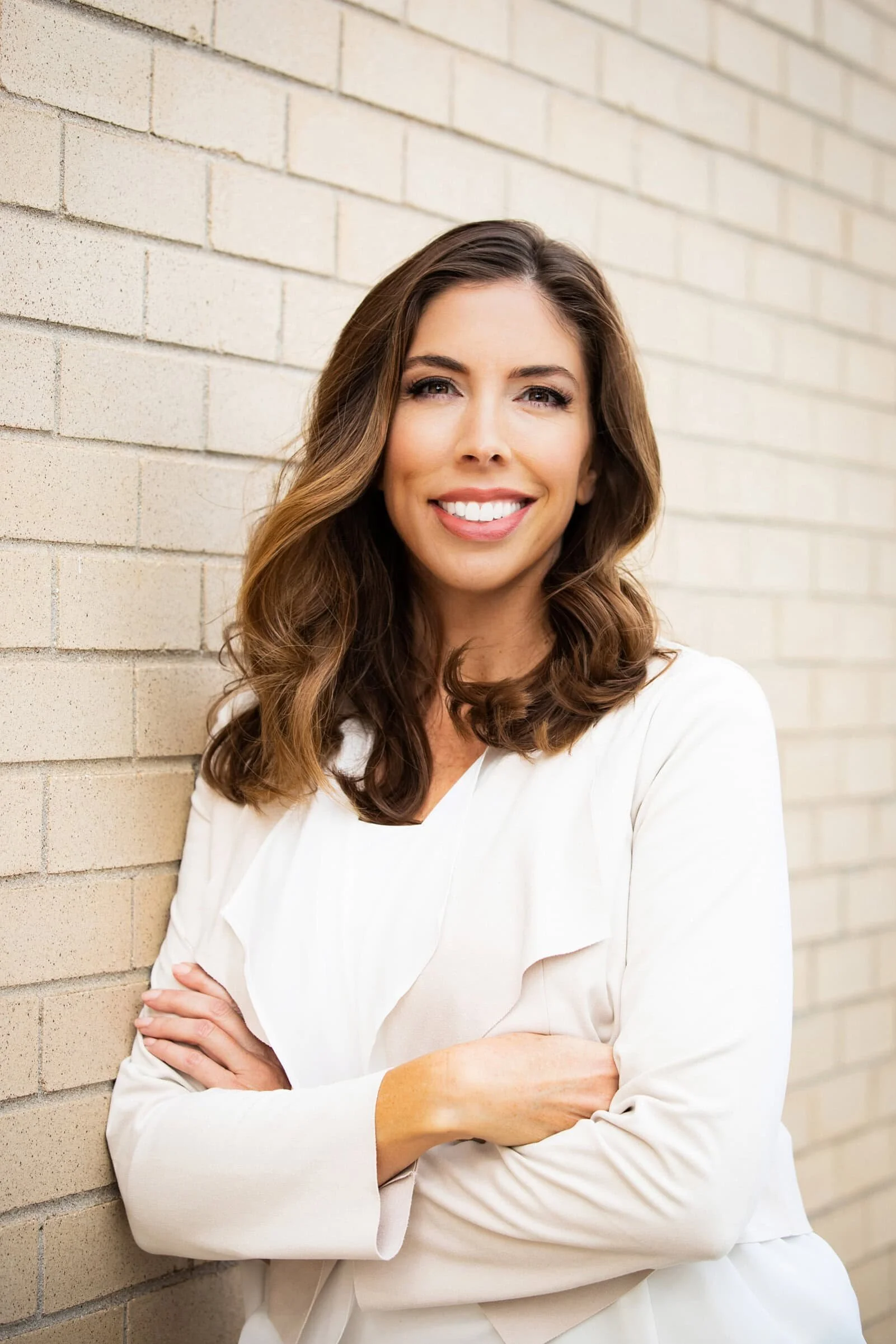 A smiling woman with wavy brown hair wearing a white blazer, standing with crossed arms against a beige brick wall.
