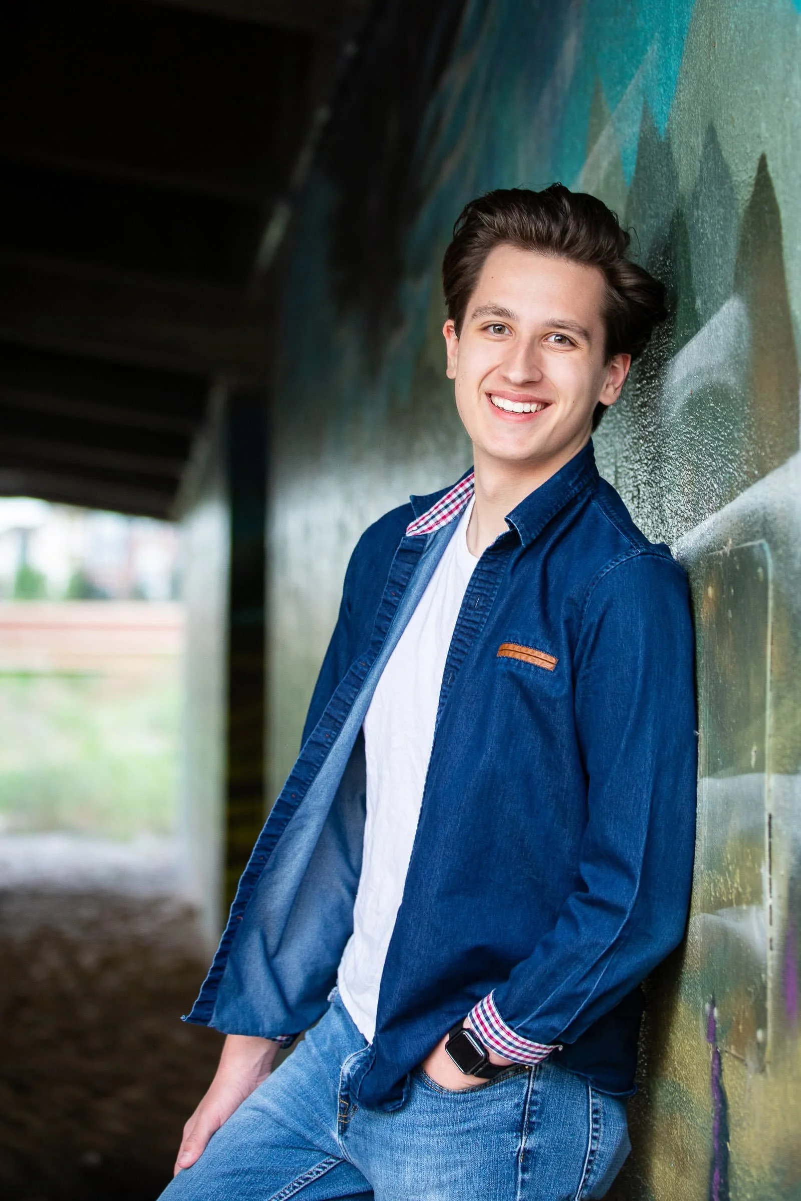 A young man with dark brown hair and fair skin smiling, leaning against a graffiti-covered wall under a bridge or overpass, wearing a denim jacket over a white t-shirt and jeans.