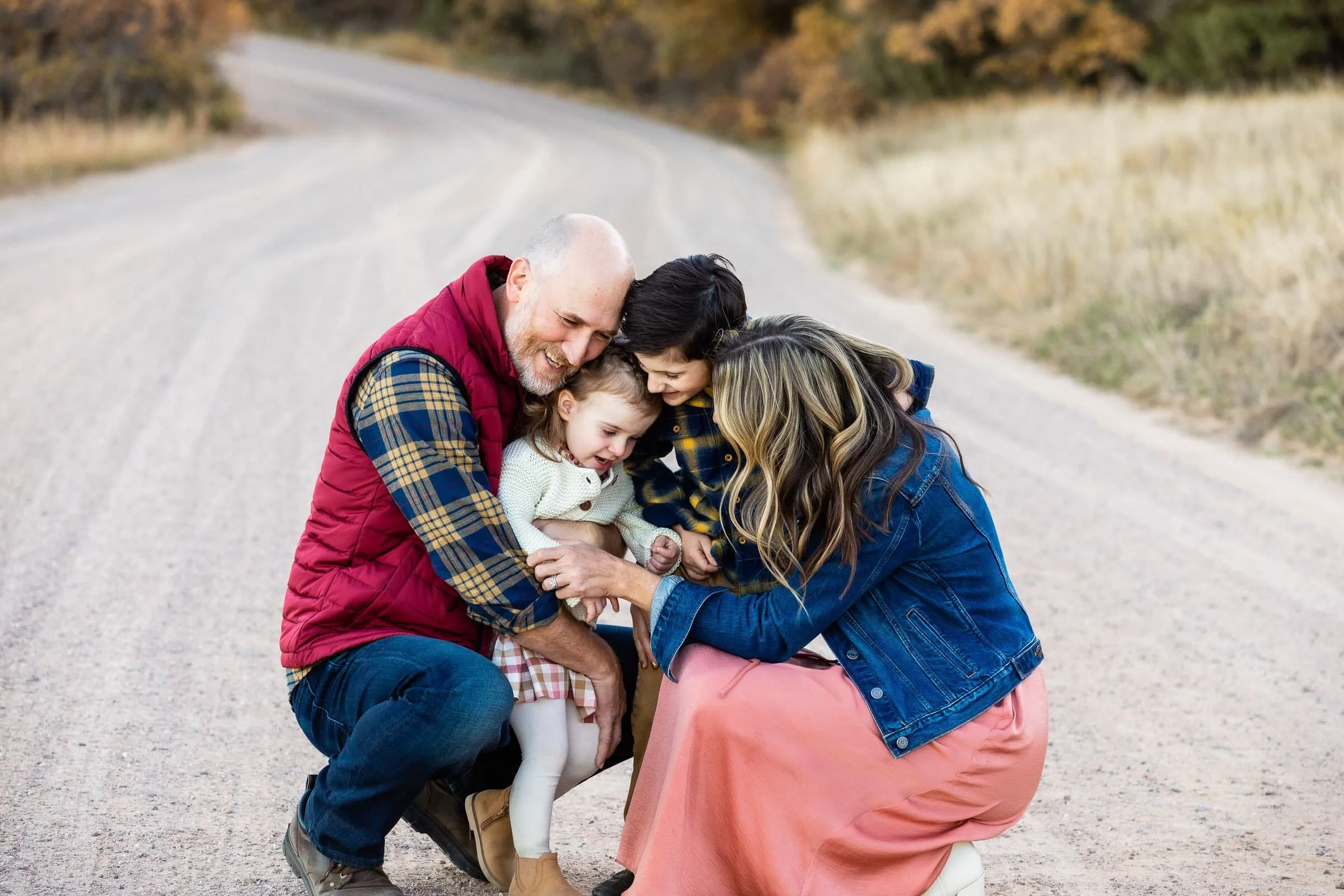 A family of four, including a father, mother, and two young daughters, are on a dirt road outdoors, all smiling and leaning closely together as they engage in an affectionate moment.