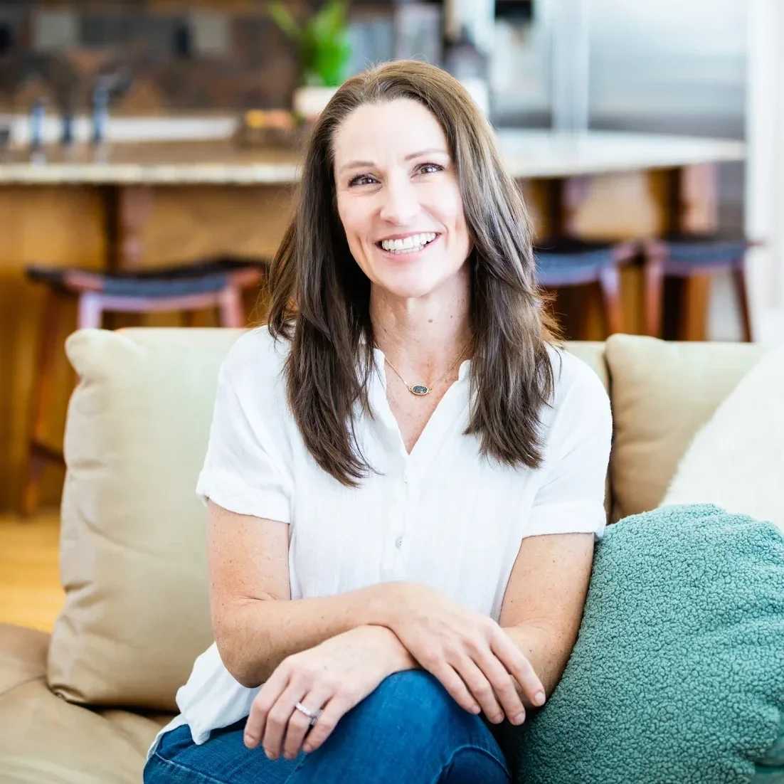 A woman with long brown hair smiling, sitting on a beige couch in a cozy, well-lit indoor space.