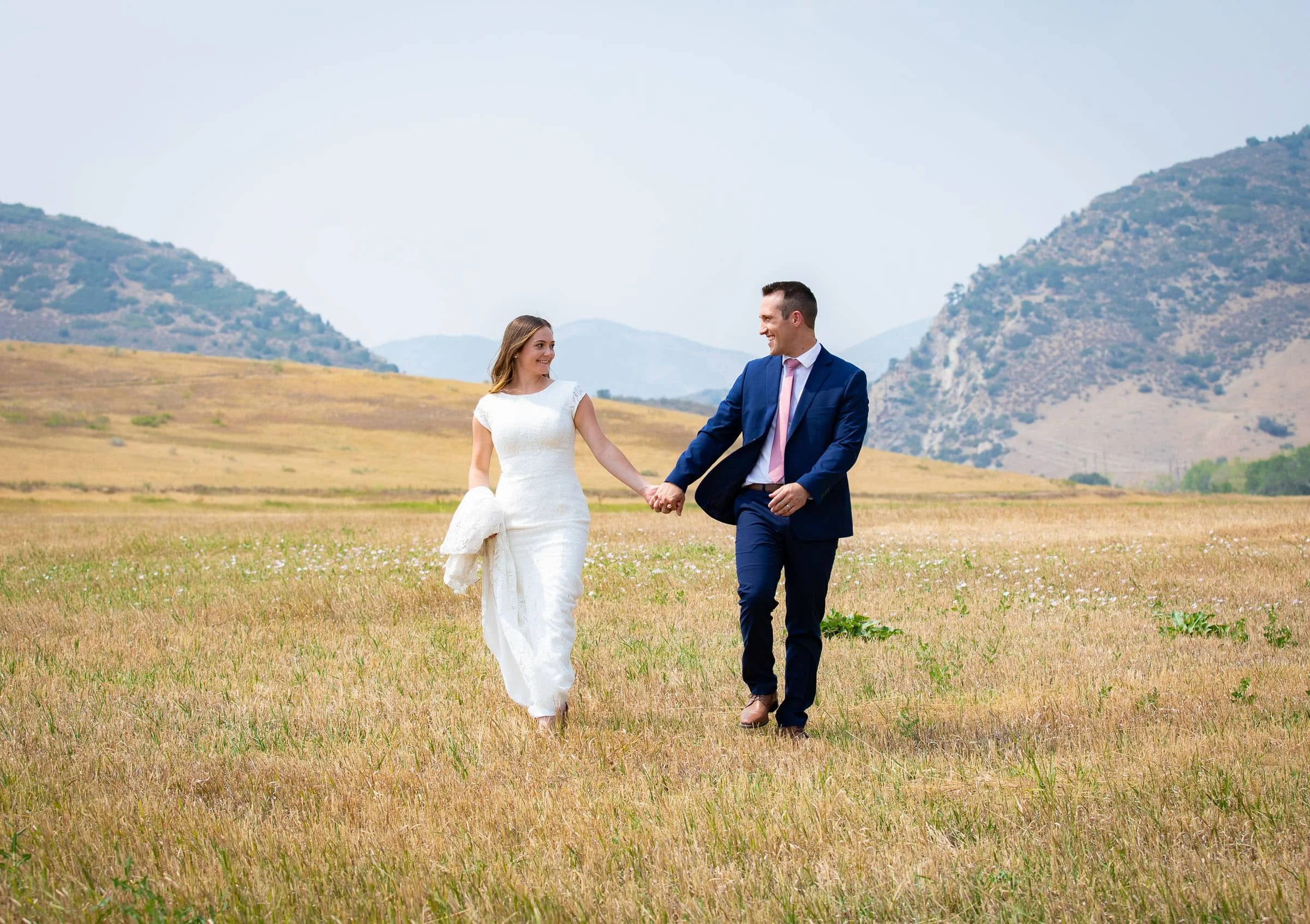 A couple walking hand in hand through a grassy field, with mountains in the background. The woman is wearing a white dress and holding her shoes and dress in one hand. The man is dressed in a blue suit with a pink tie.