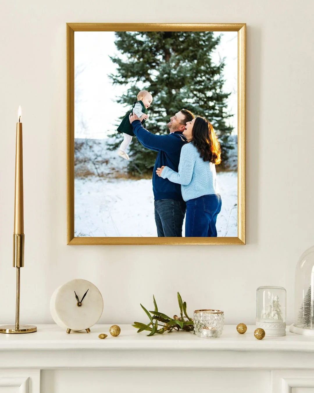 A family photo of two parents and a baby outdoors in winter with snow and a large pine tree, viewed through a window.