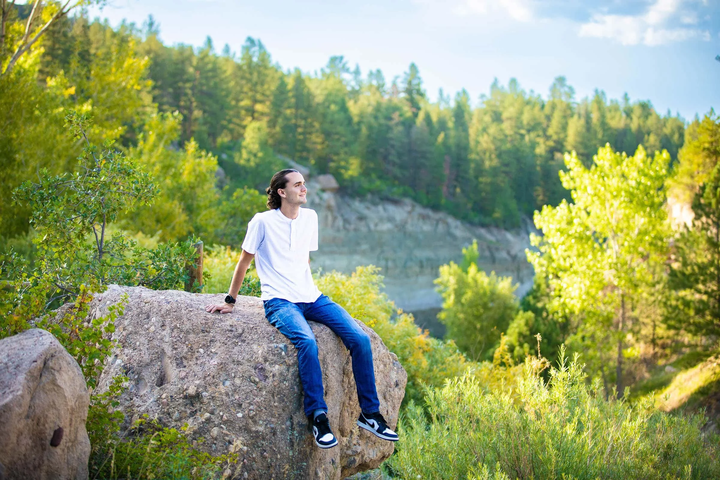 A young man in a white shirt and jeans sitting on a large rock outdoors surrounded by green trees and foliage, with a scenic forested background.
