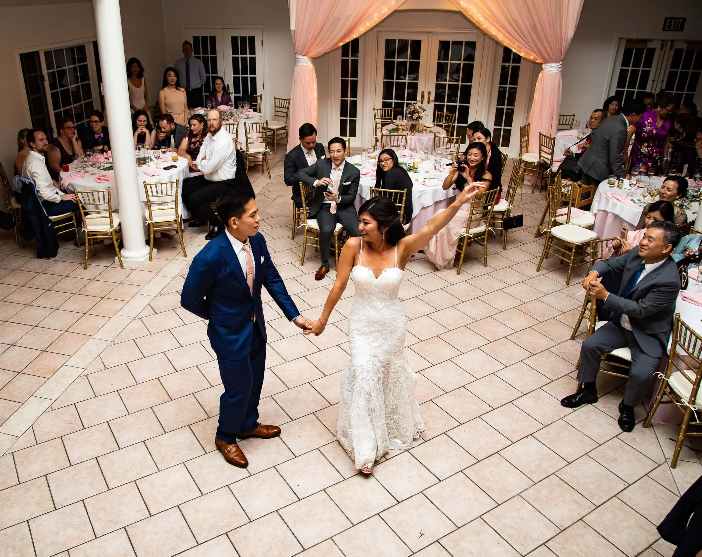 A bride and groom are dancing at their wedding reception, holding hands, with wedding guests seated at round tables watching and taking photos inside a decorated banquet hall.