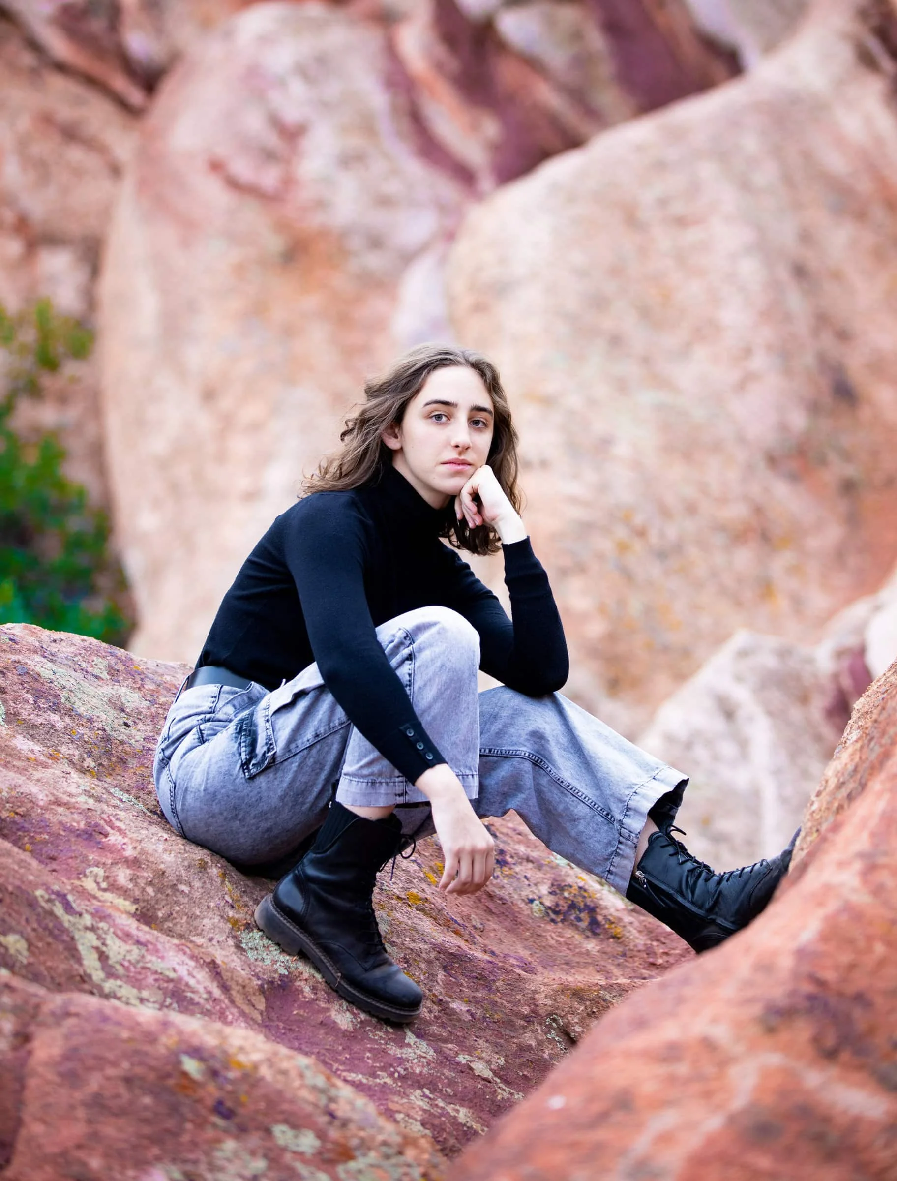 A woman sitting on a large, reddish-pink rock formation outdoors, wearing a black turtleneck, baggy light gray jeans, and black lace-up boots, with her hand resting on her chin and looking at the camera.