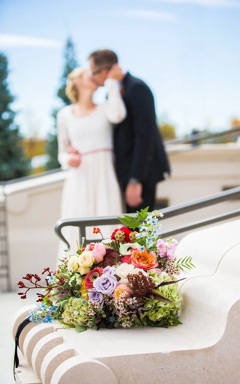 A colorful wedding bouquet of various flowers resting on a white bench. In the background, a blurred couple, likely the bride and groom, sharing a kiss.