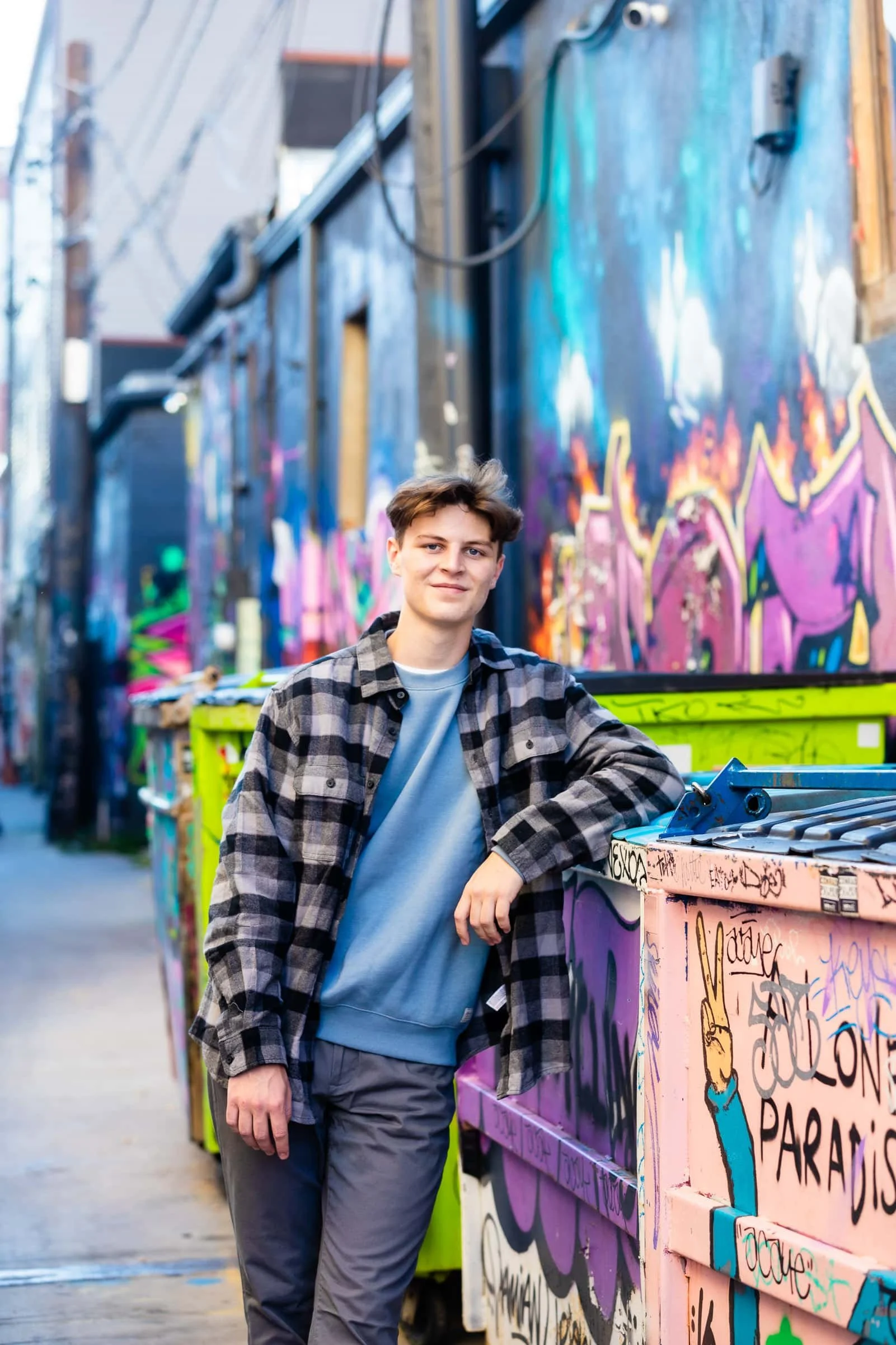 A young man with short brown hair, wearing a black and gray plaid shirt over a gray sweatshirt, standing next to brightly colored graffiti-covered walls and a graffiti-decorated dumpster, with a city alley background.