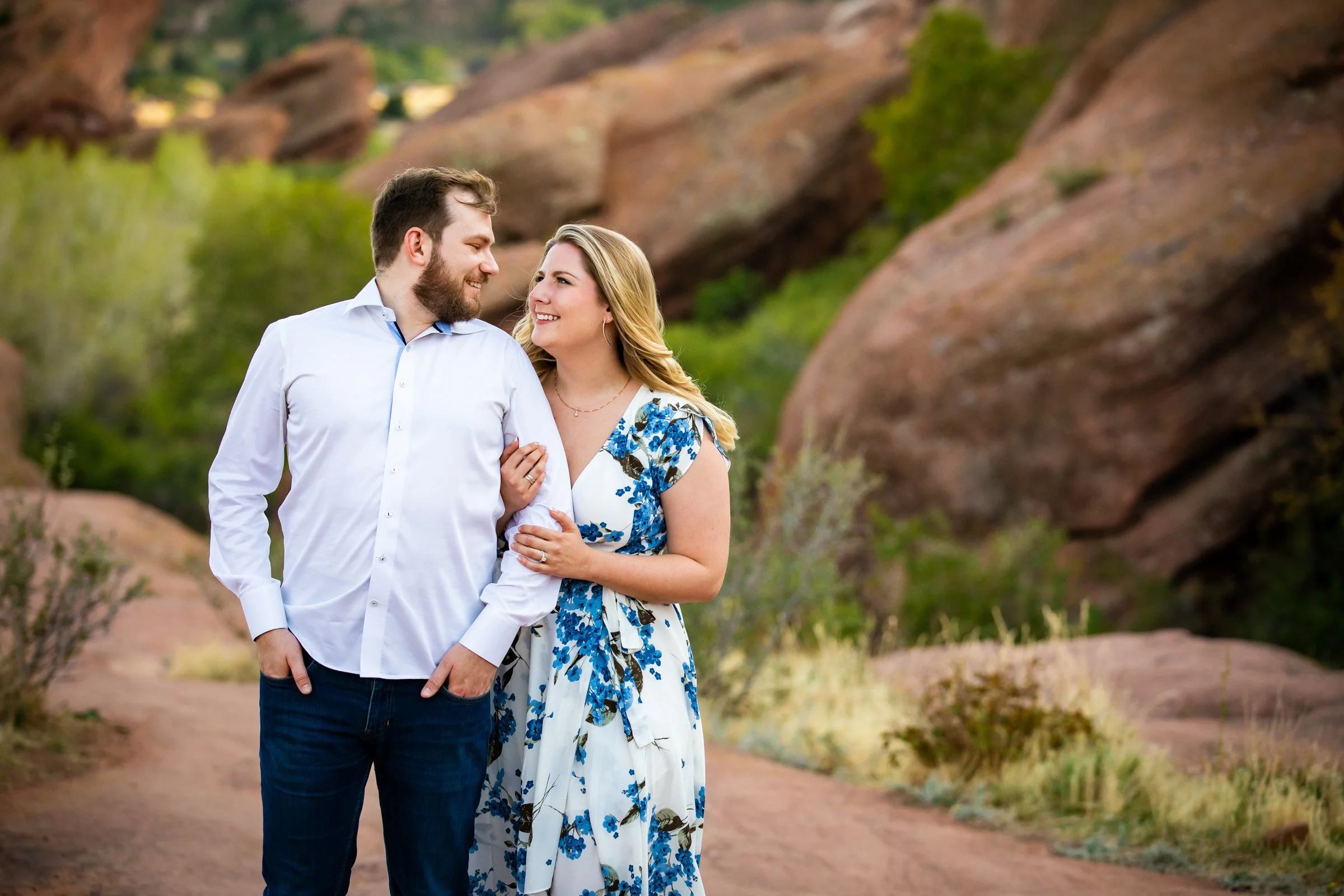 A happy couple walking in a desert landscape with large rocks and green bushes, with the woman holding the man's arm and looking at him.