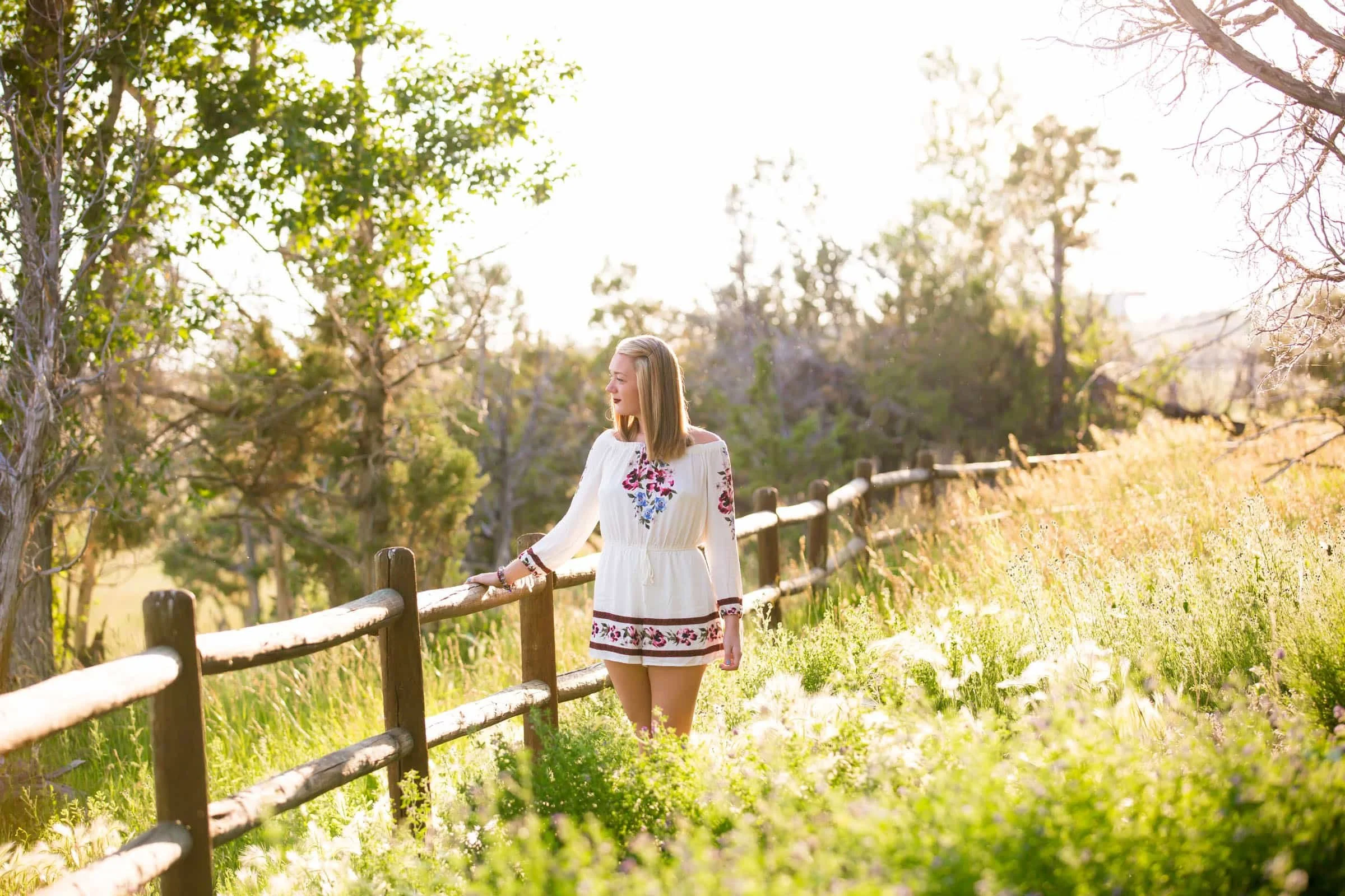 A young woman with blonde hair wearing a white embroidered dress standing by a wooden fence in a sunlit meadow with trees and greenery.