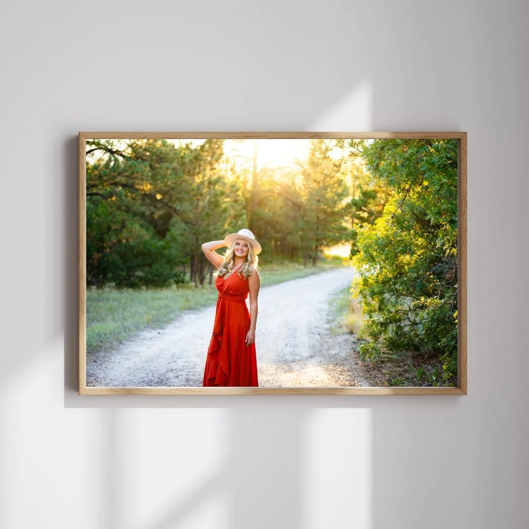 A woman in a red dress and wide-brimmed hat standing on a dirt path surrounded by trees, with sunlight shining through the leaves.