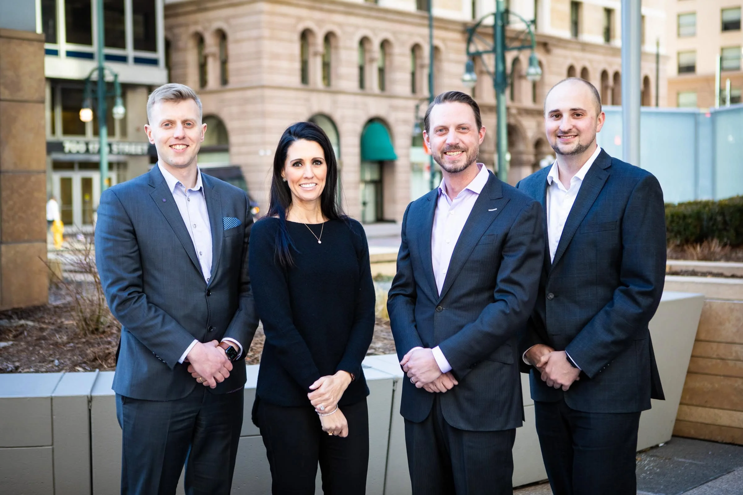 Four professionally dressed people standing outside in an urban setting with tall buildings in the background.