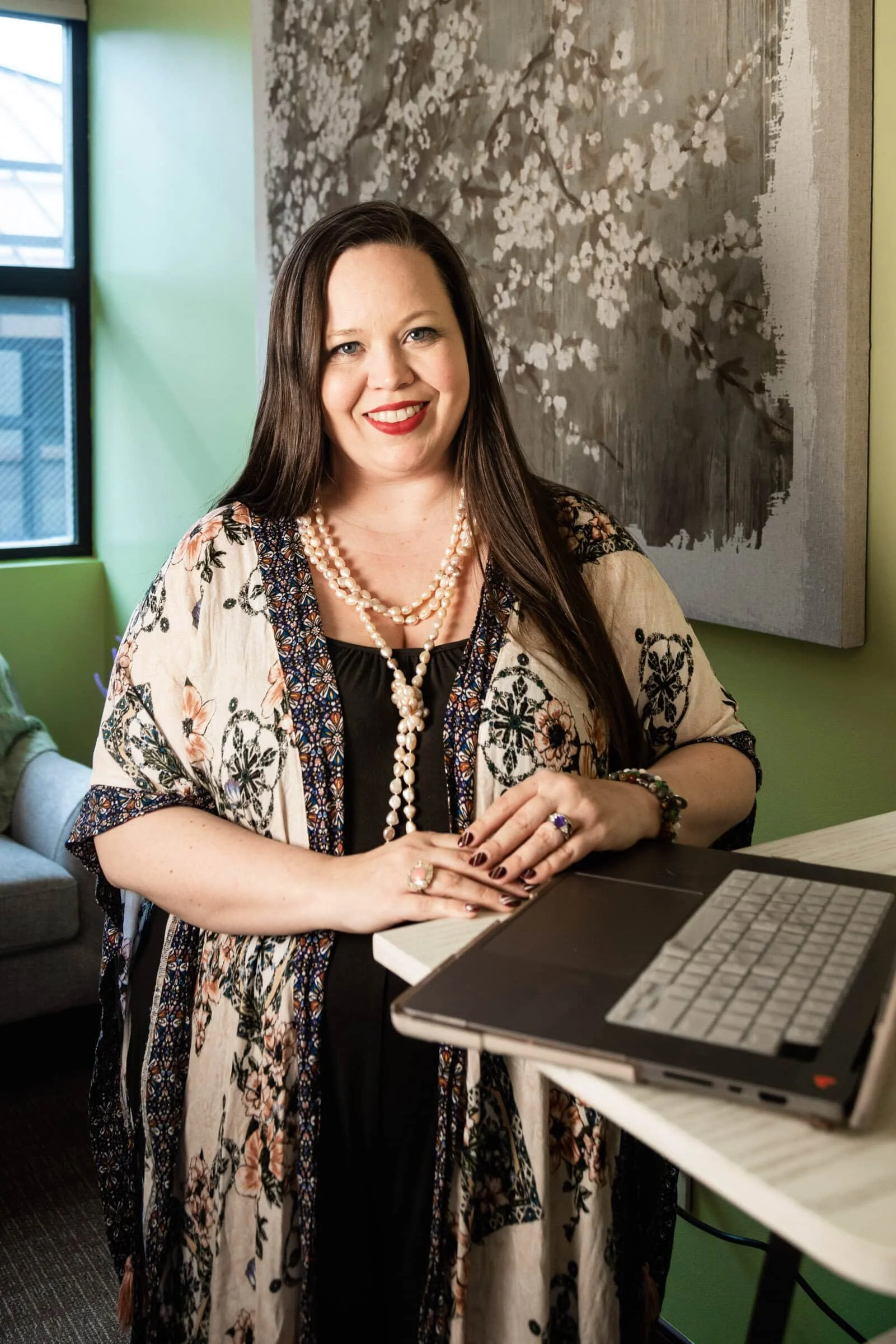 A woman with long dark hair dressed in a floral and black outfit, wearing pearl necklaces and rings, standing next to a computer on a white table, in a room with green walls and a large abstract painting.
