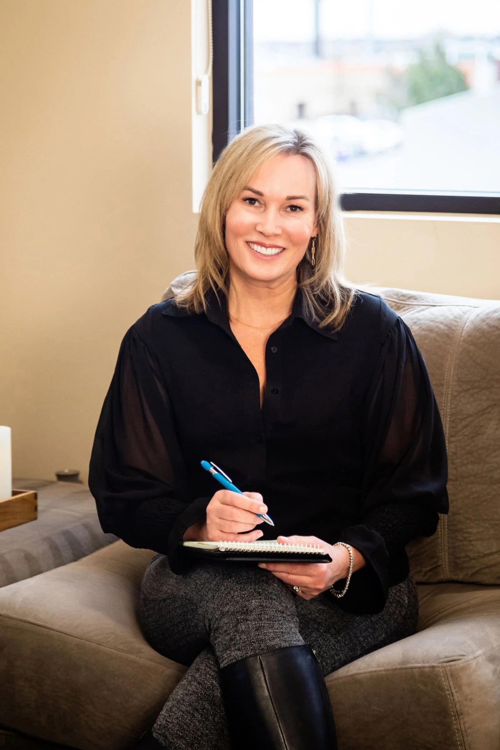 A smiling woman with blonde hair sitting on a beige sofa near a window, holding a blue pen and a notebook.