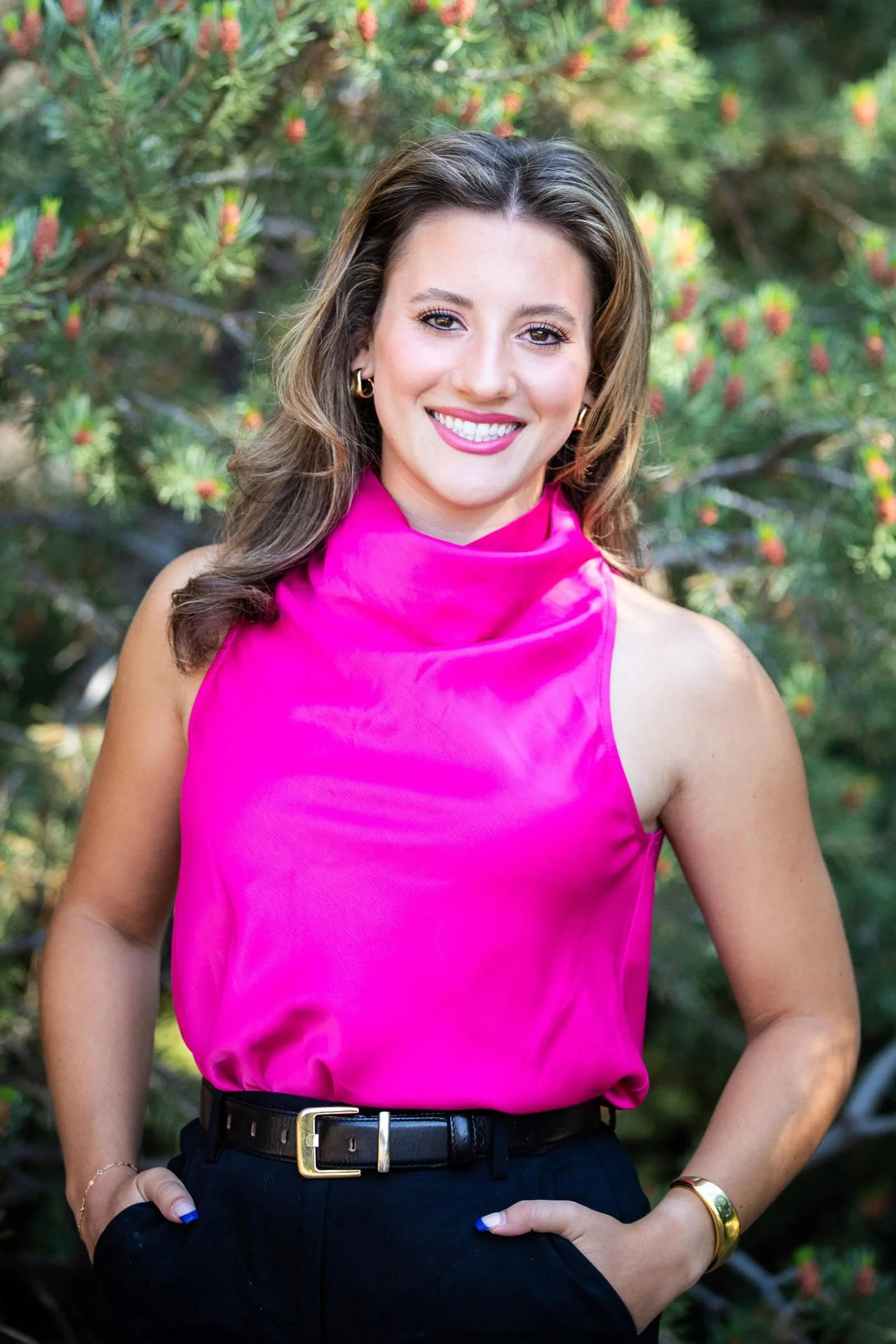 A woman with brown hair wearing a bright pink sleeveless top, black pants, and gold accessories, standing outdoors in front of green foliage.