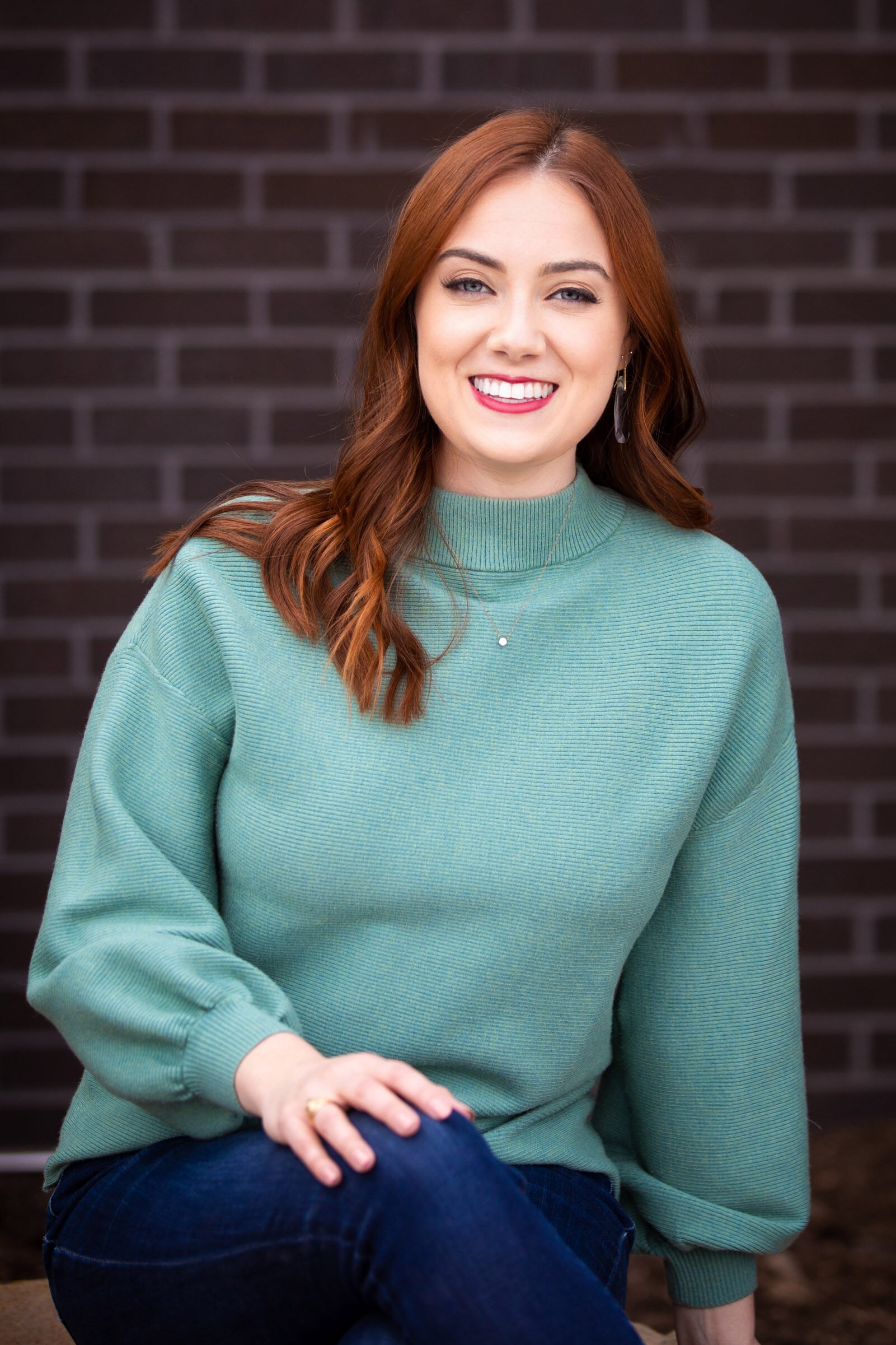 A young woman with red hair, wearing a green sweater and blue jeans, smiling while sitting against a dark brick wall.
