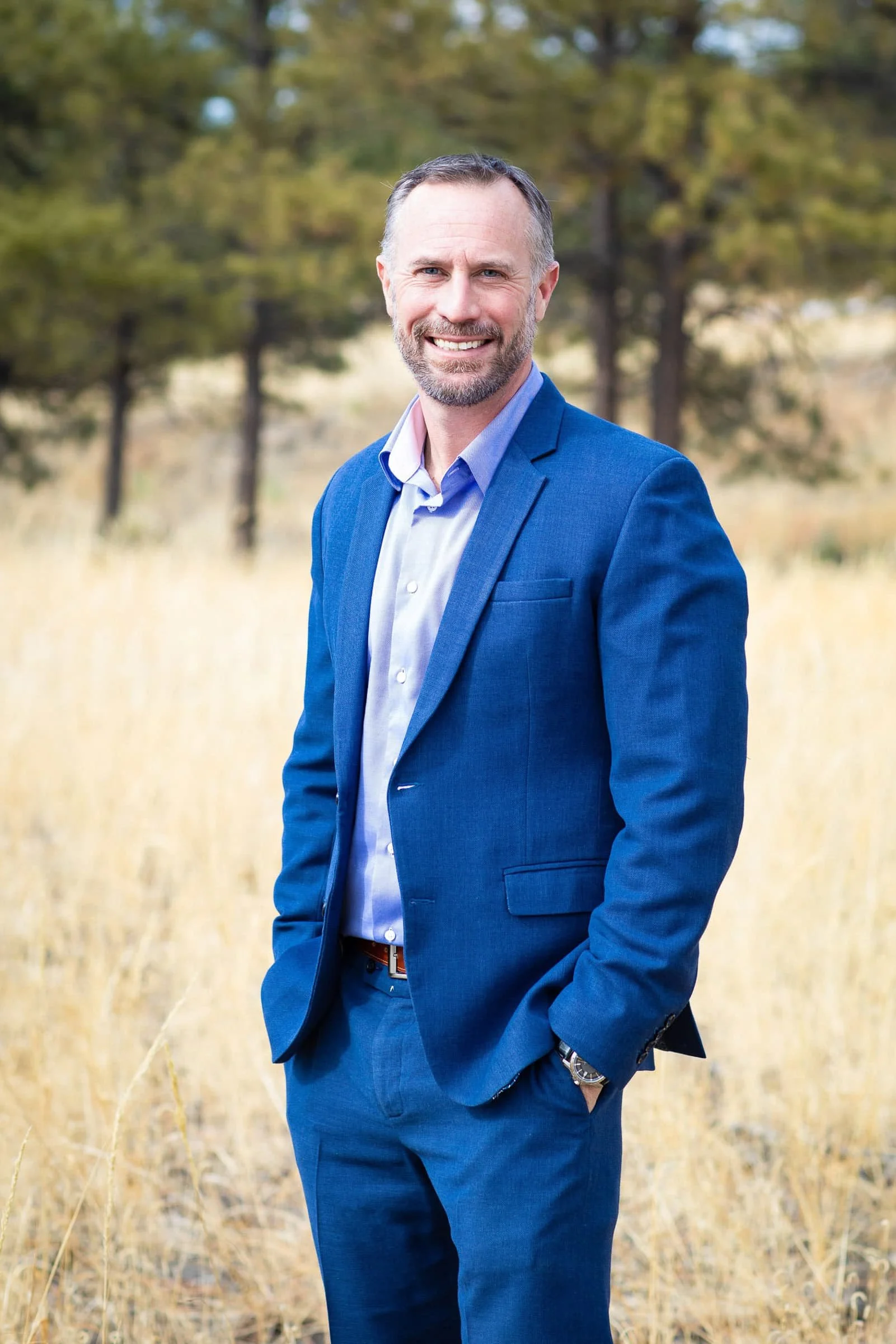A man in a blue suit standing in a field with yellow grass and trees in the background.
