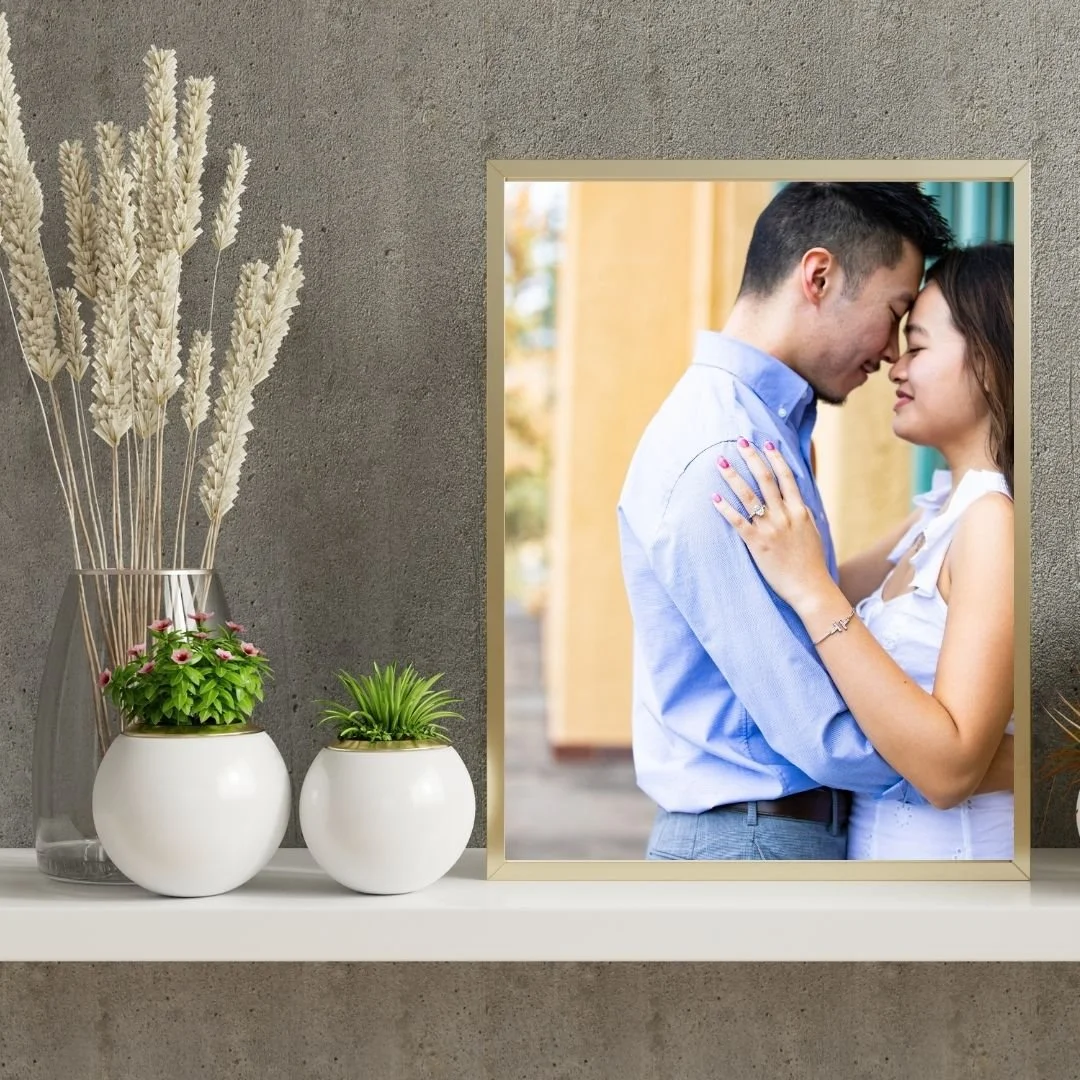 A framed photo of a couple touching foreheads, standing close with eyes closed, on a shelf beside potted plants and dried wheat in a vase in front of a gray wall.
