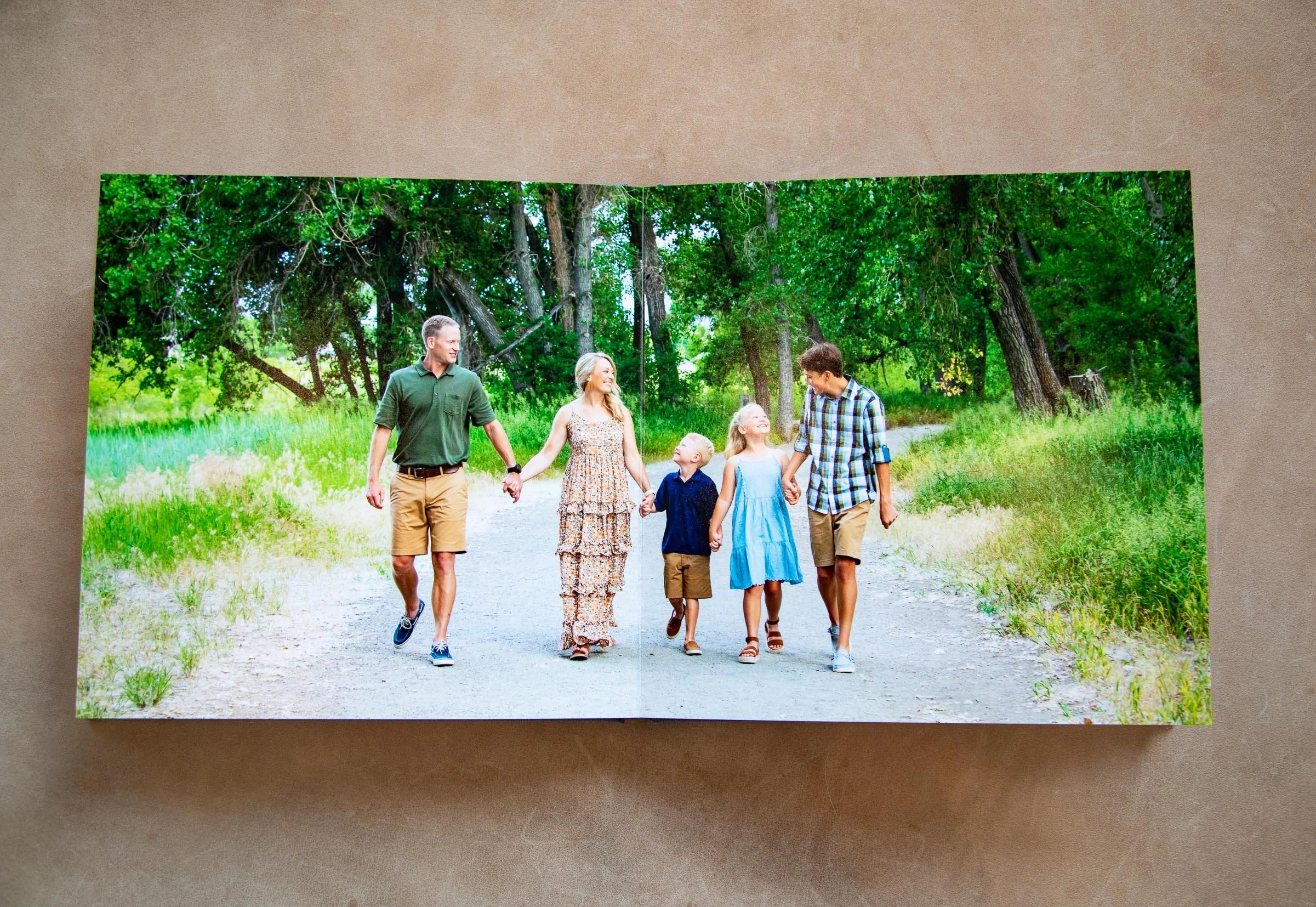 A family of six walking together on a dirt path in a green park. The family consists of two adult men, two young girls, a woman, and a young boy, all holding hands and enjoying a sunny day.