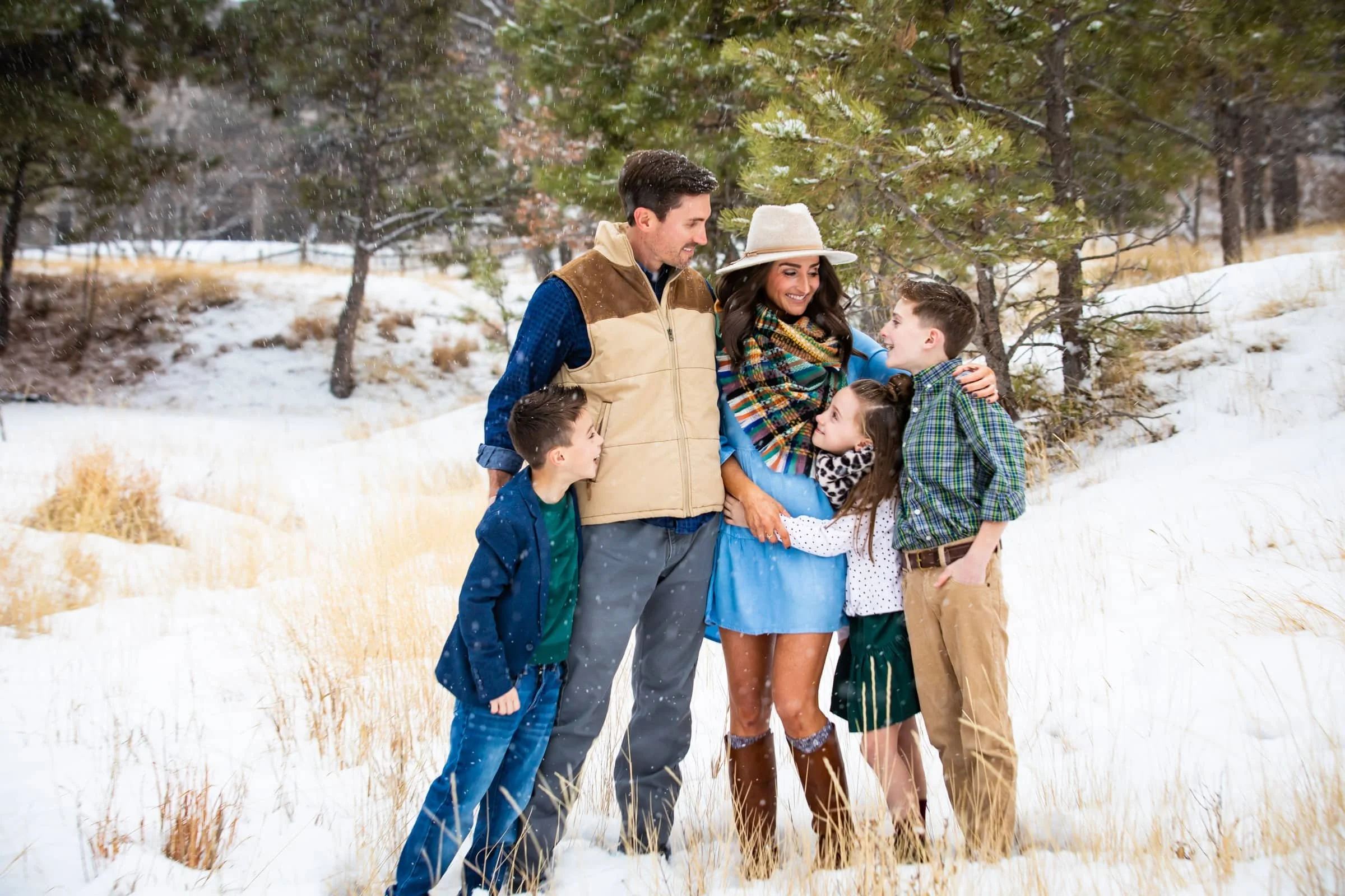 Family of six enjoying winter outdoors in snowy landscape with trees, smiling and embracing.