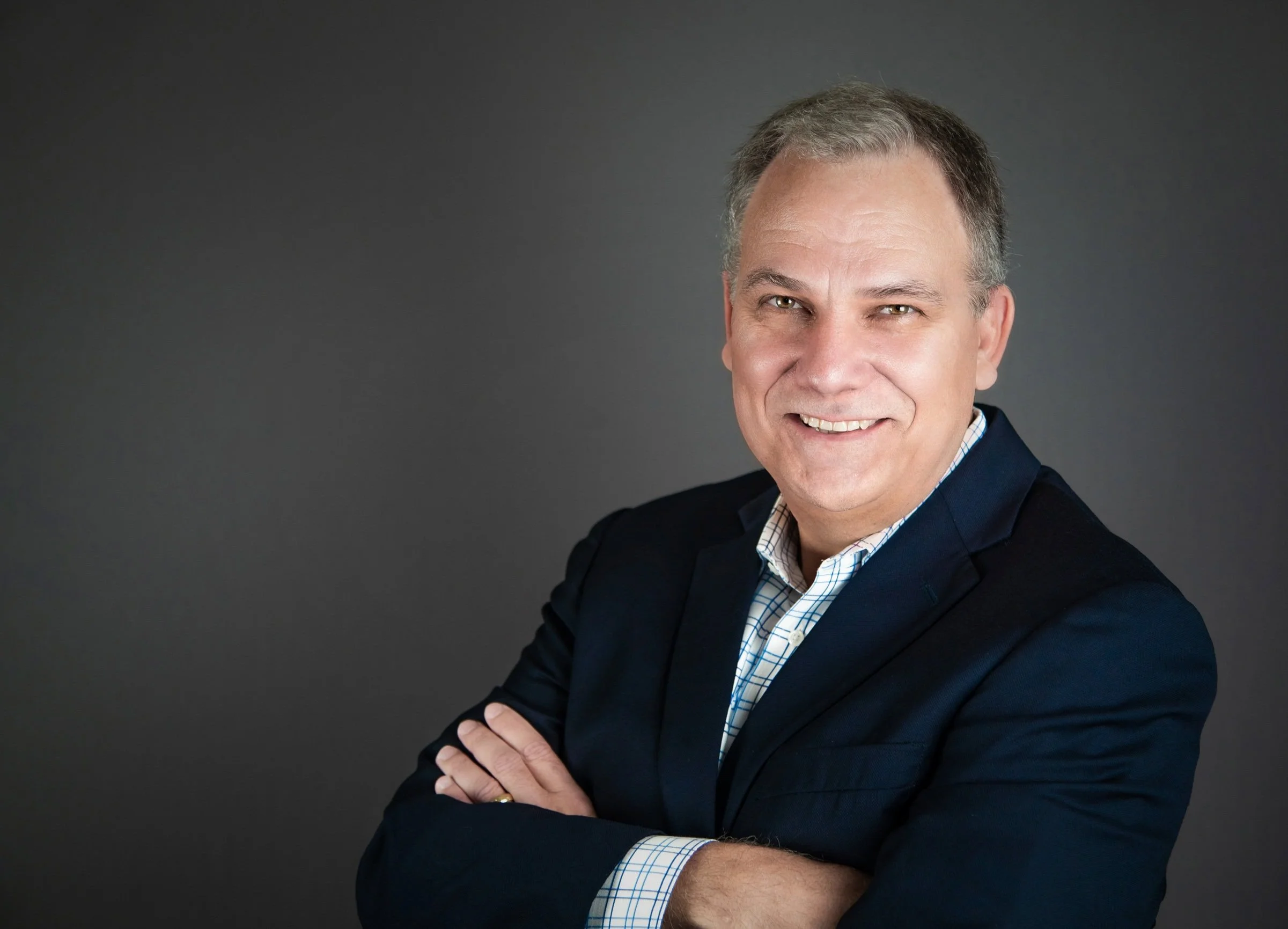 A middle-aged man with gray hair smiling, wearing a dark blazer over a plaid shirt, standing with arms crossed against a dark gray background.