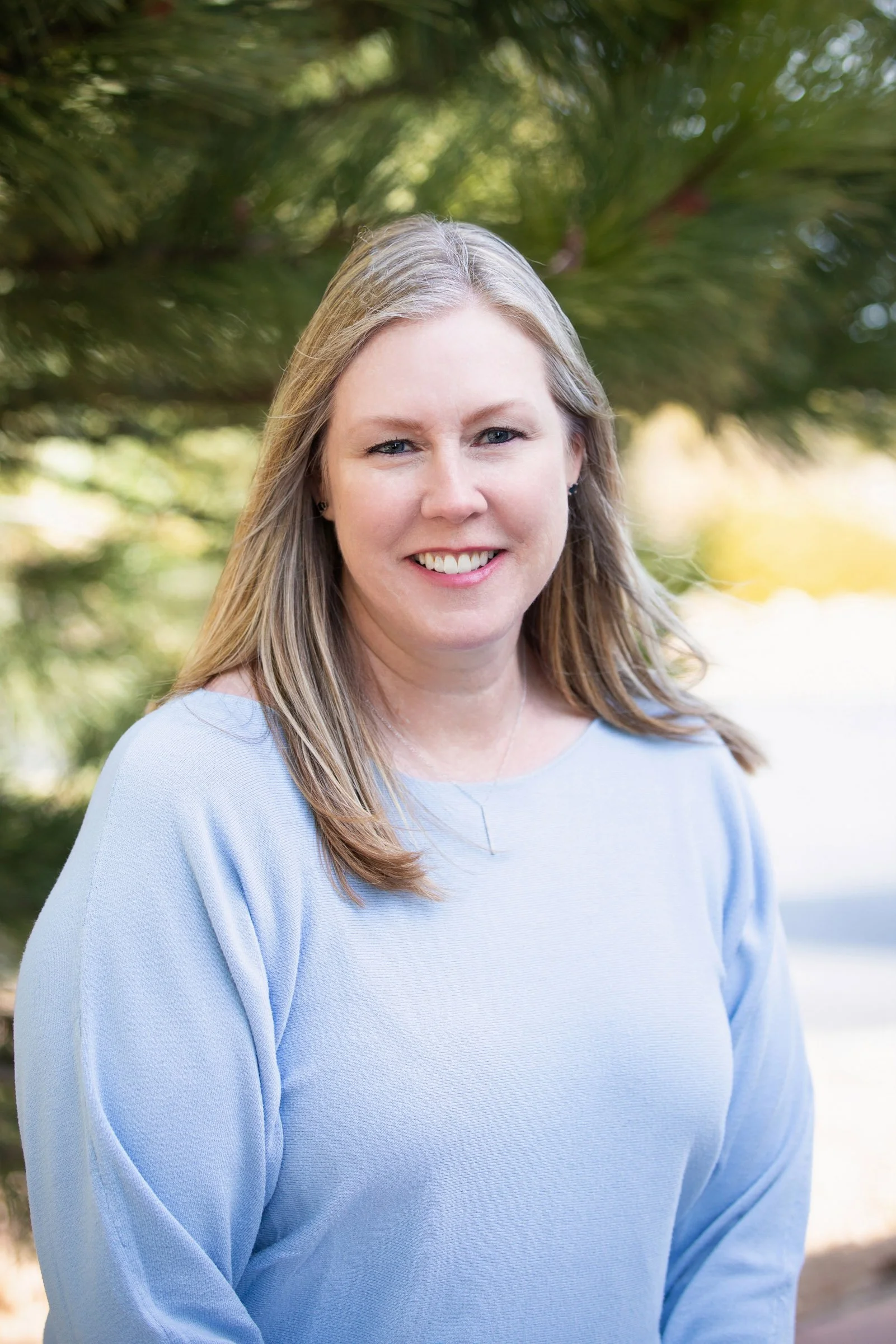 A woman with light brown hair and blue eyes smiling outdoors, wearing a light blue top, with a background of green trees and sunlight.