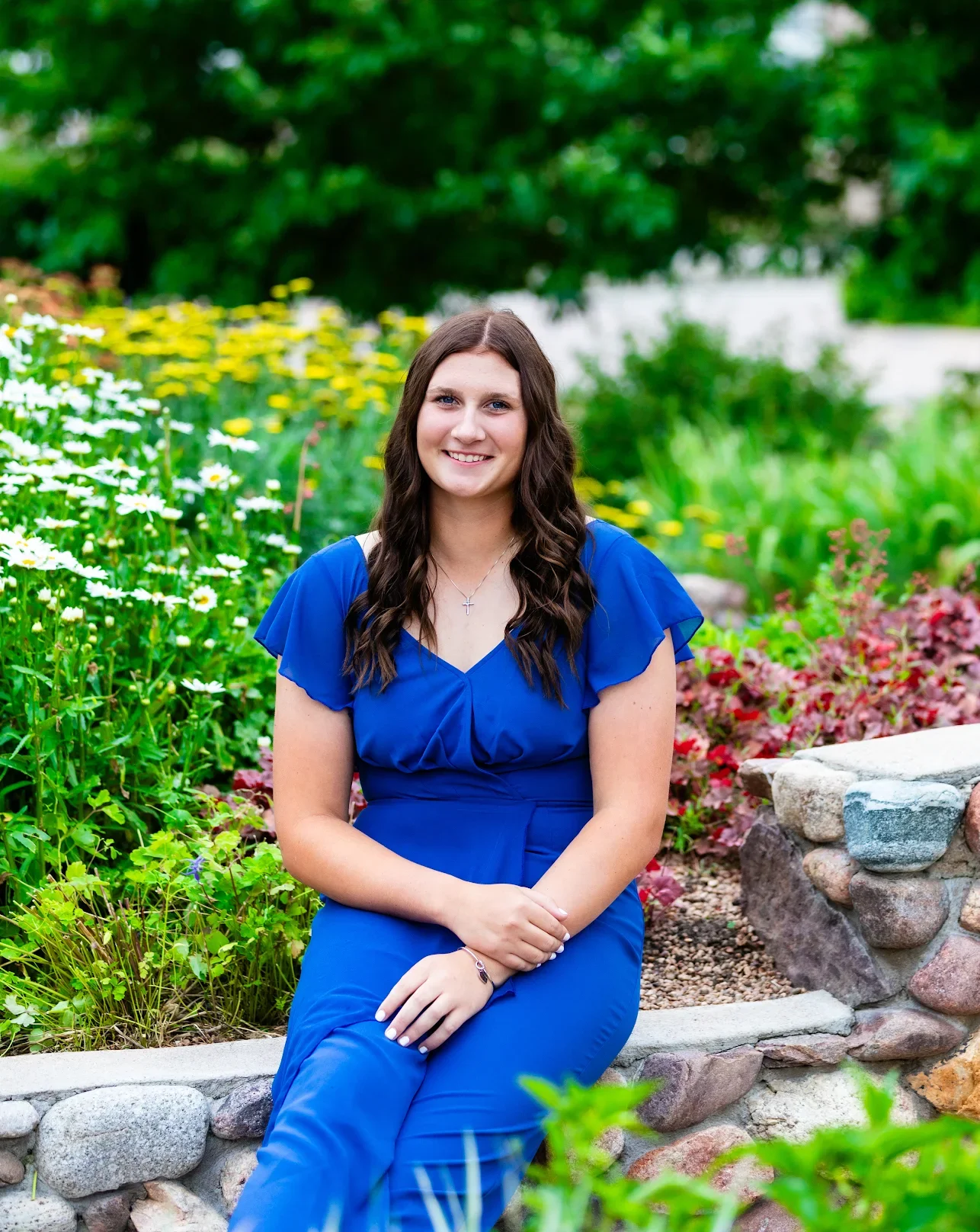 A young woman with long brown hair in a blue dress, sitting on a stone border in a garden with green trees and colorful flowers.