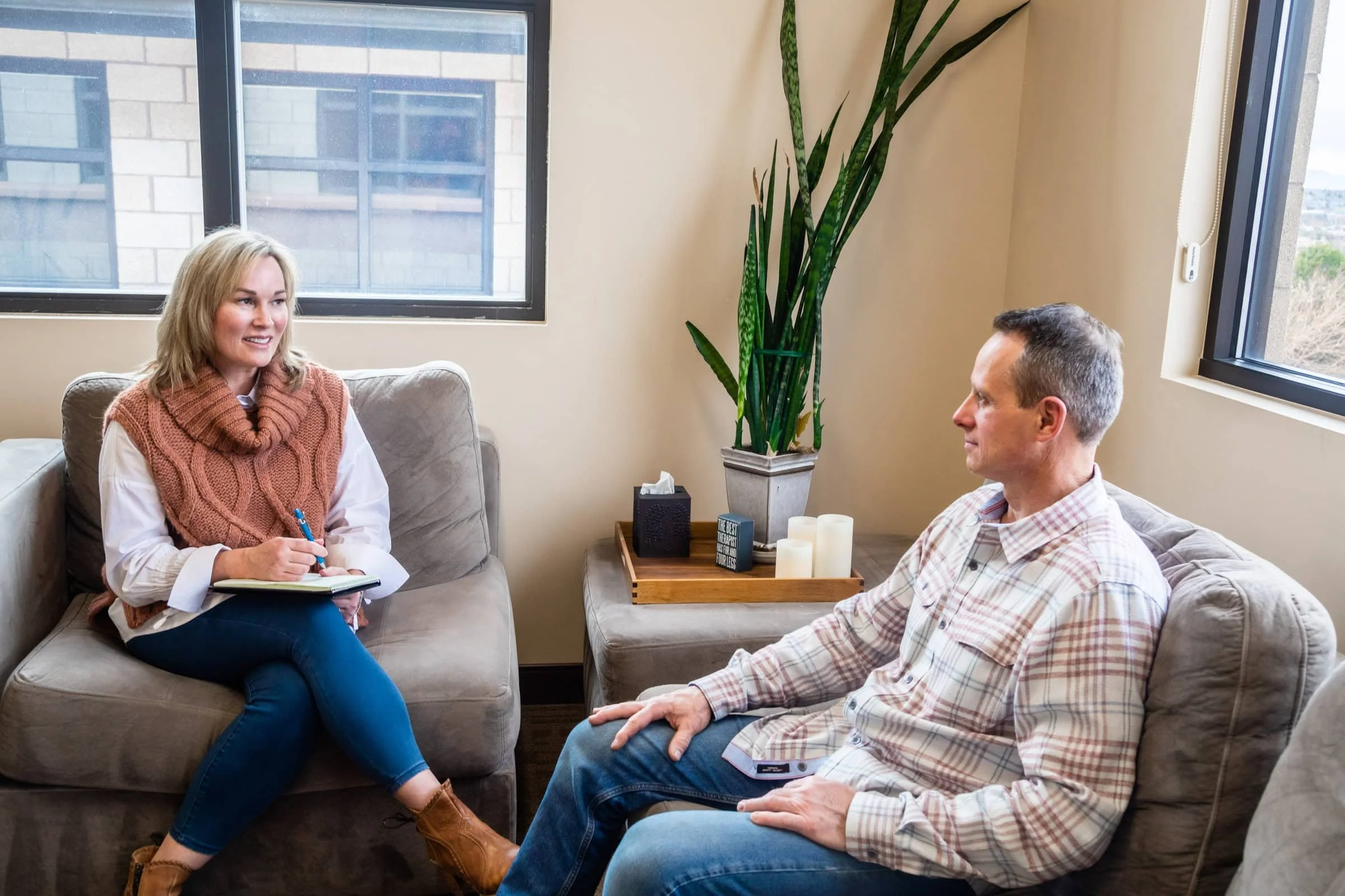 A woman and a man are sitting on sofas in a therapy or counseling session. The woman is holding a notepad and pen, smiling while talking to the man. The room has large windows, a tall green plant, and a tray with candles on a side table.