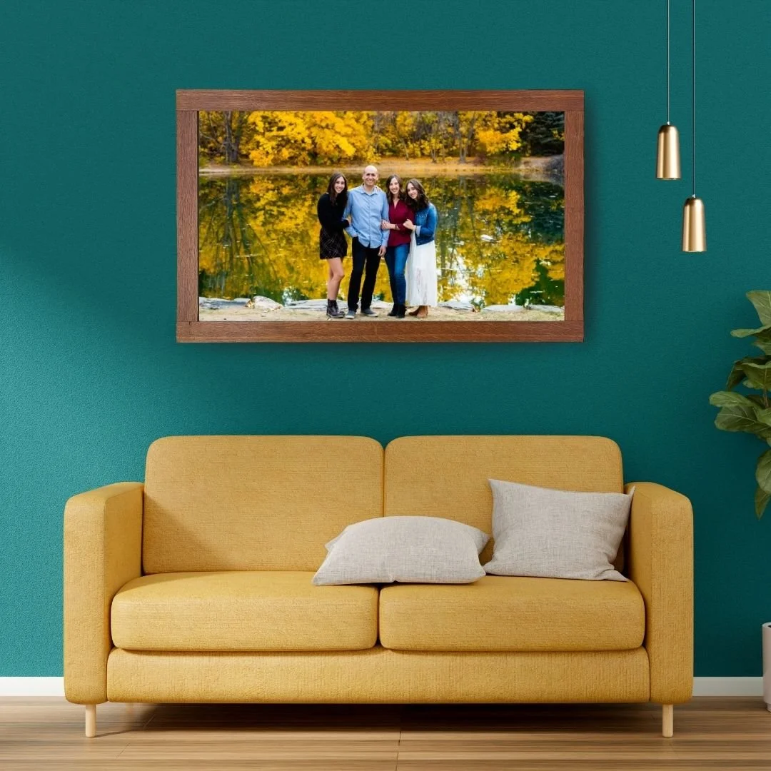 A framed photograph of four people standing by a lake with yellow autumn trees in the background, hanging on a teal wall above a yellow sofa with two beige pillows.