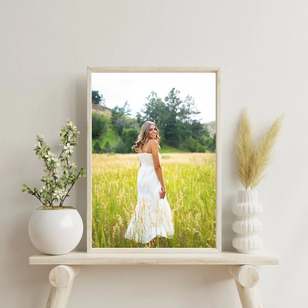 Framed photo of a woman in a white dress standing in a grassy field with trees and mountains in the background, displayed on a wooden shelf with white vases containing flowers and pampas grass.