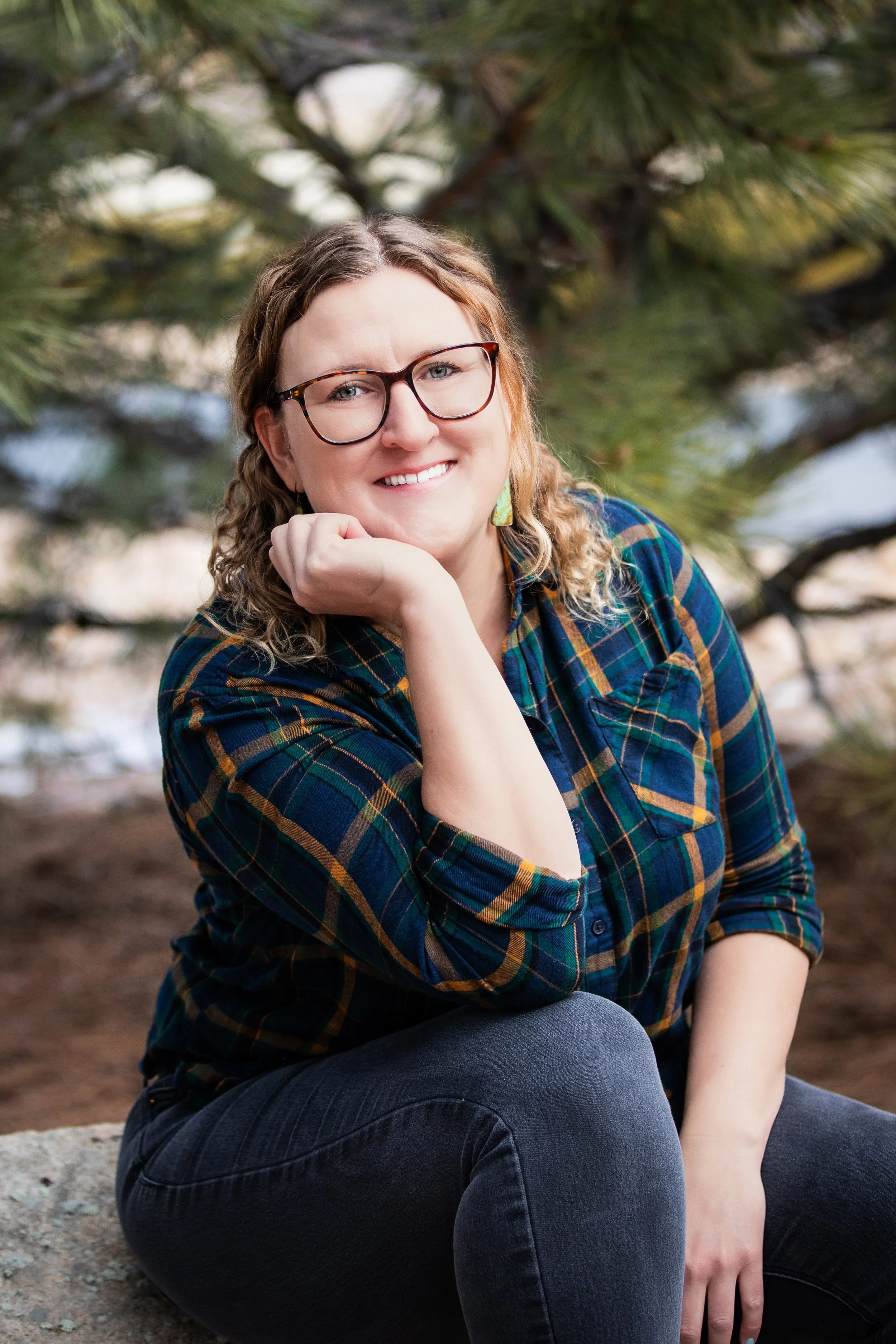 A woman with curly blonde hair, glasses, and a plaid shirt sitting outdoors near a pine tree, smiling at the camera.
