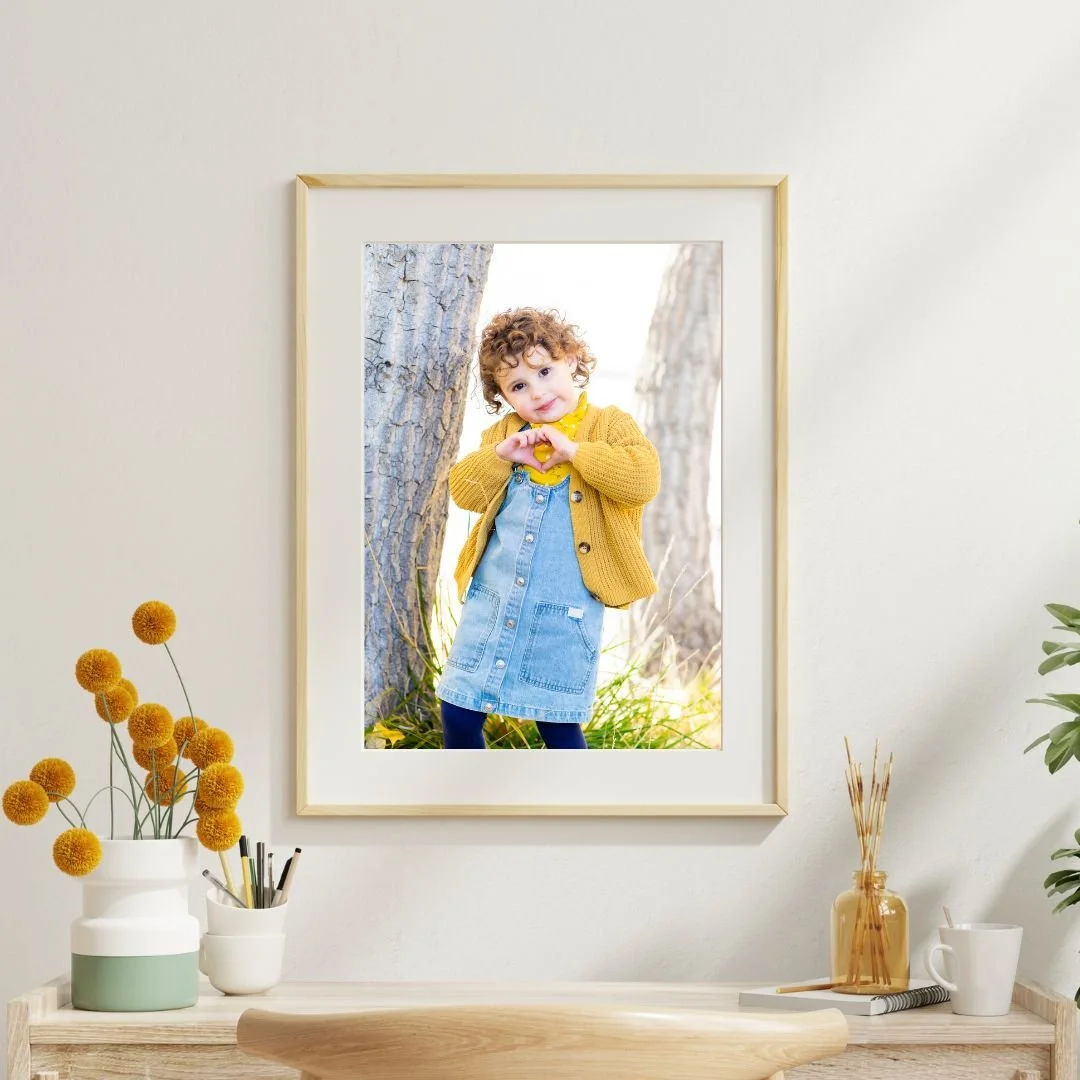 Interior wall with a framed photo of a young girl with curly hair, wearing a yellow sweater, denim dress, and dark tights, standing outdoors near trees, making a heart shape with her hands.