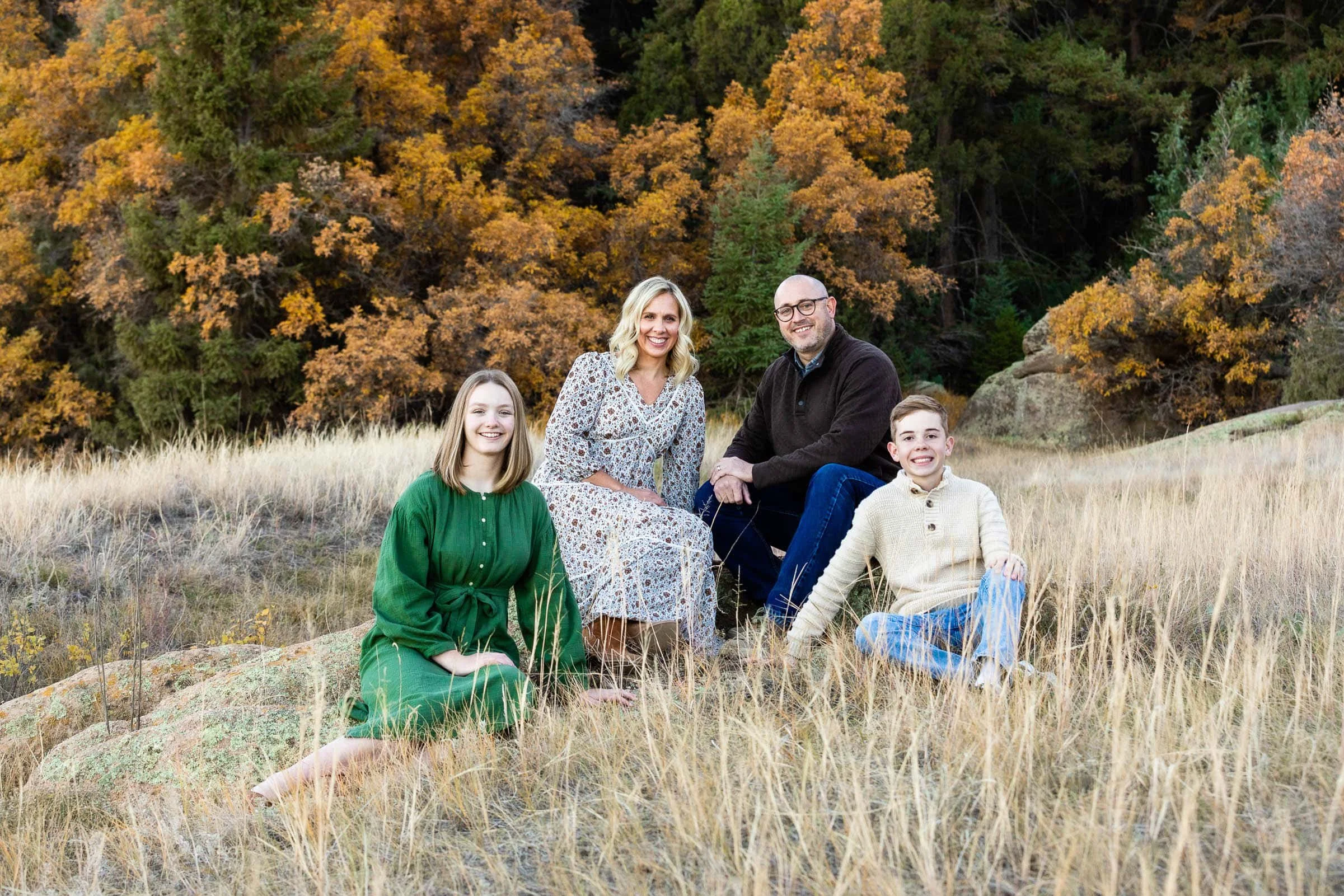 Family of four sitting on a grassy hillside with fall foliage and trees in the background.