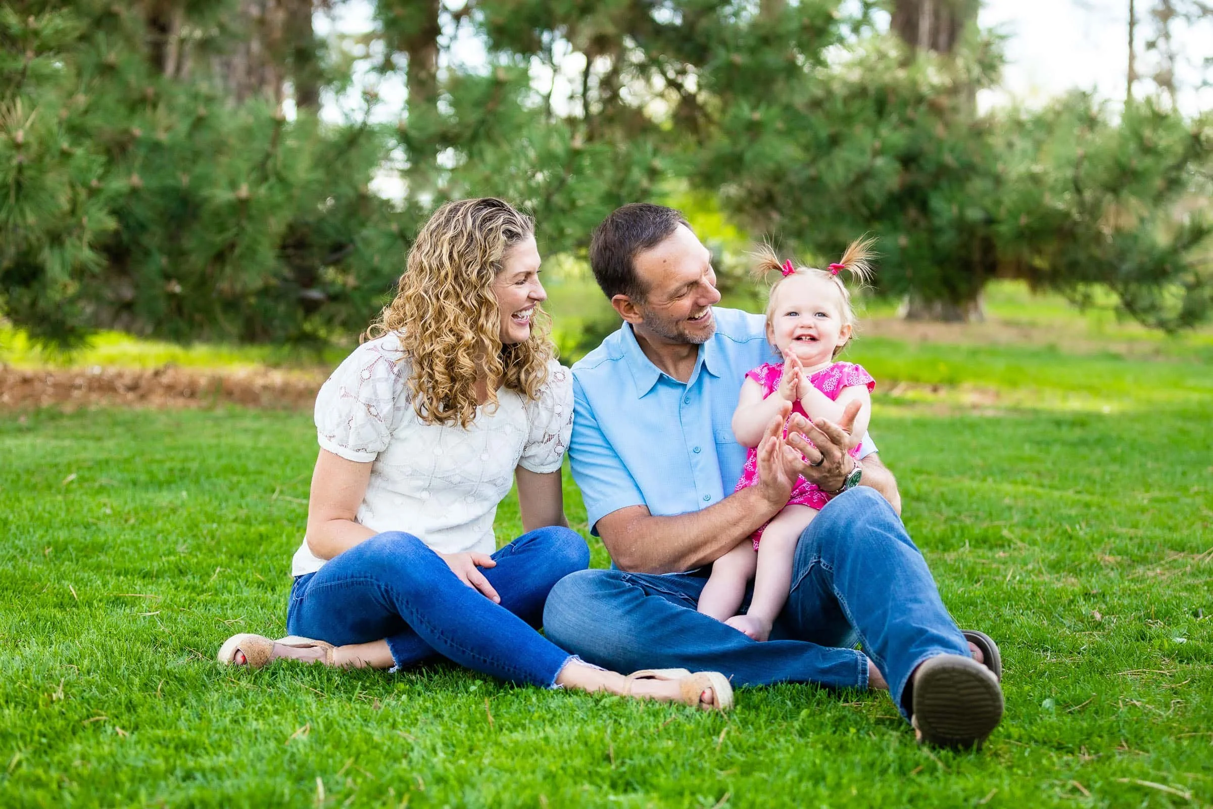 Family sitting on grass in a park, laughing and enjoying time together with a toddler girl in a pink dress.