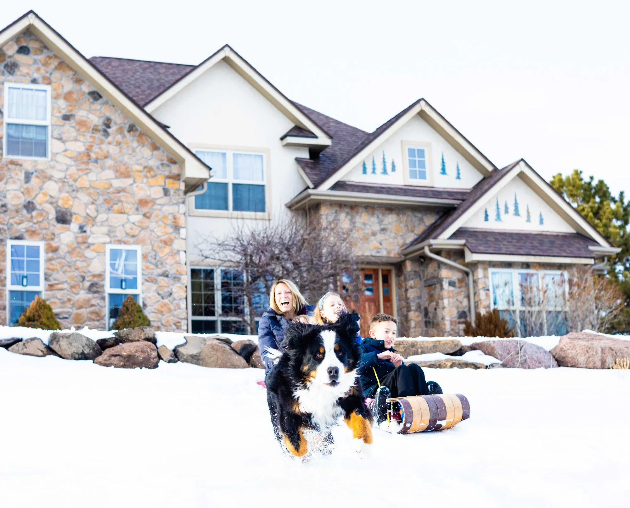 A family of three, including a woman, a young girl, and a boy, with a black and white dog, enjoying sledding on snow in front of a large stone house with multiple roofs and windows, during winter.