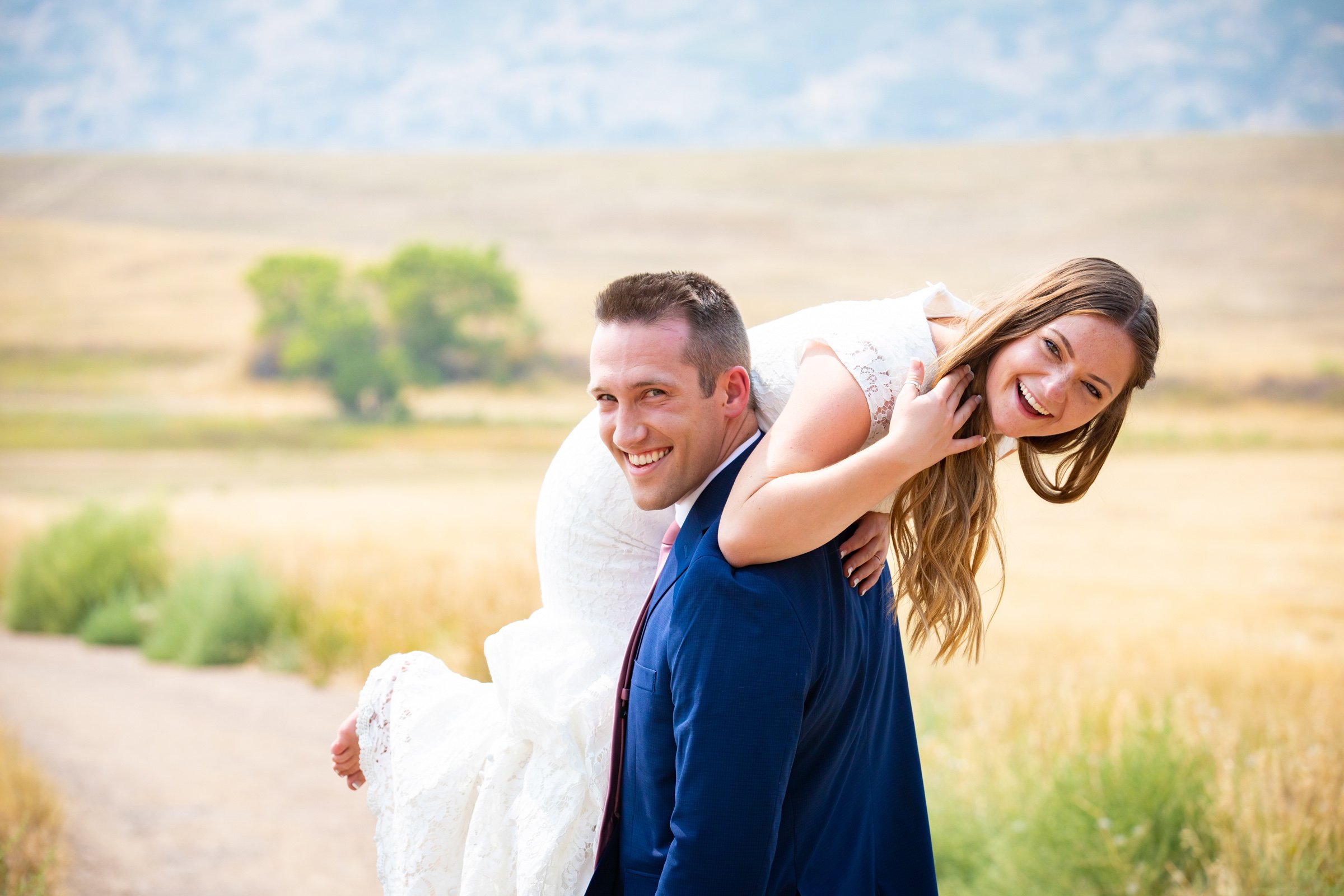 A couple outdoors, with the man carrying the woman on his back, both smiling and laughing in a rural landscape with grassy fields and distant mountains.