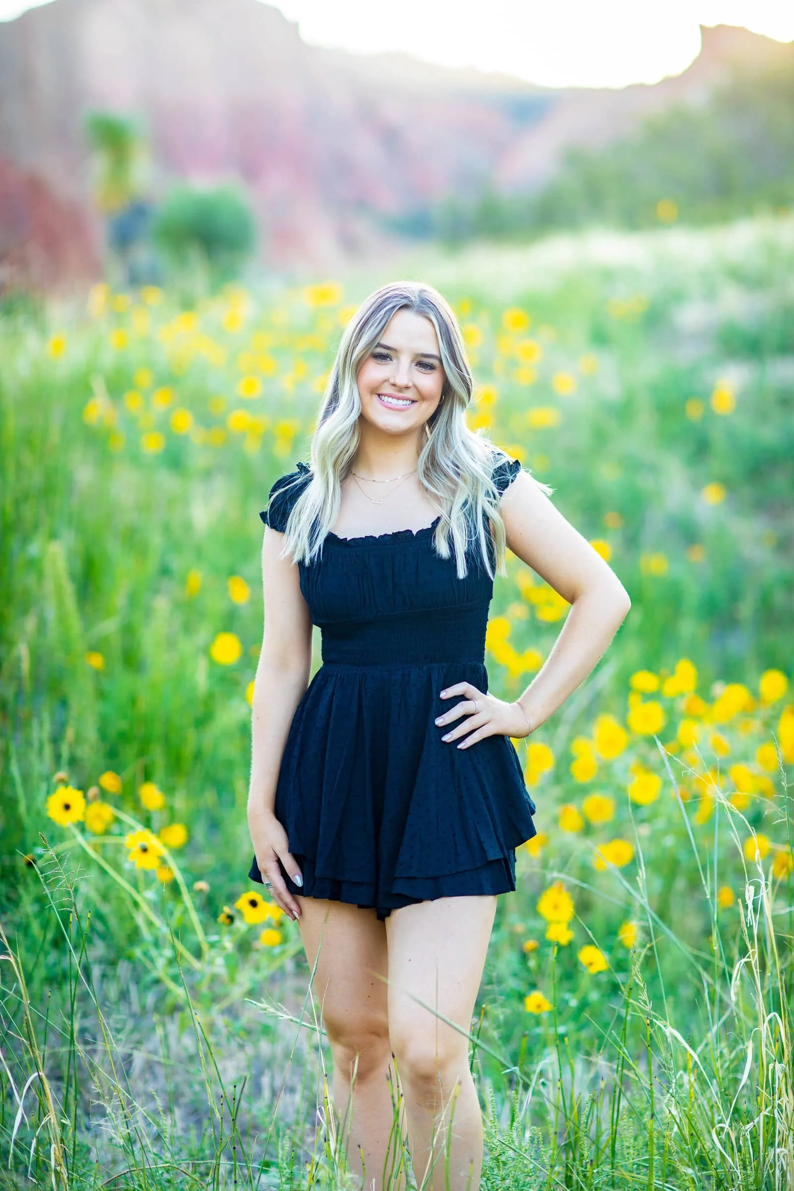 A young woman in a black dress standing in a field of yellow flowers with a blurred background of green hills and trees.