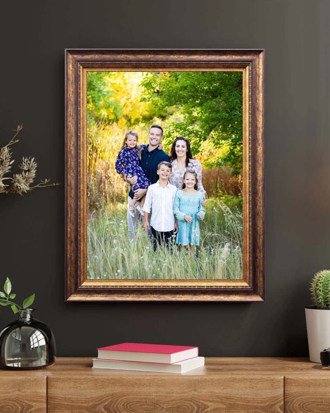Framed family portrait of two adults and three children standing in a grassy field with dense green and yellow trees in the background.