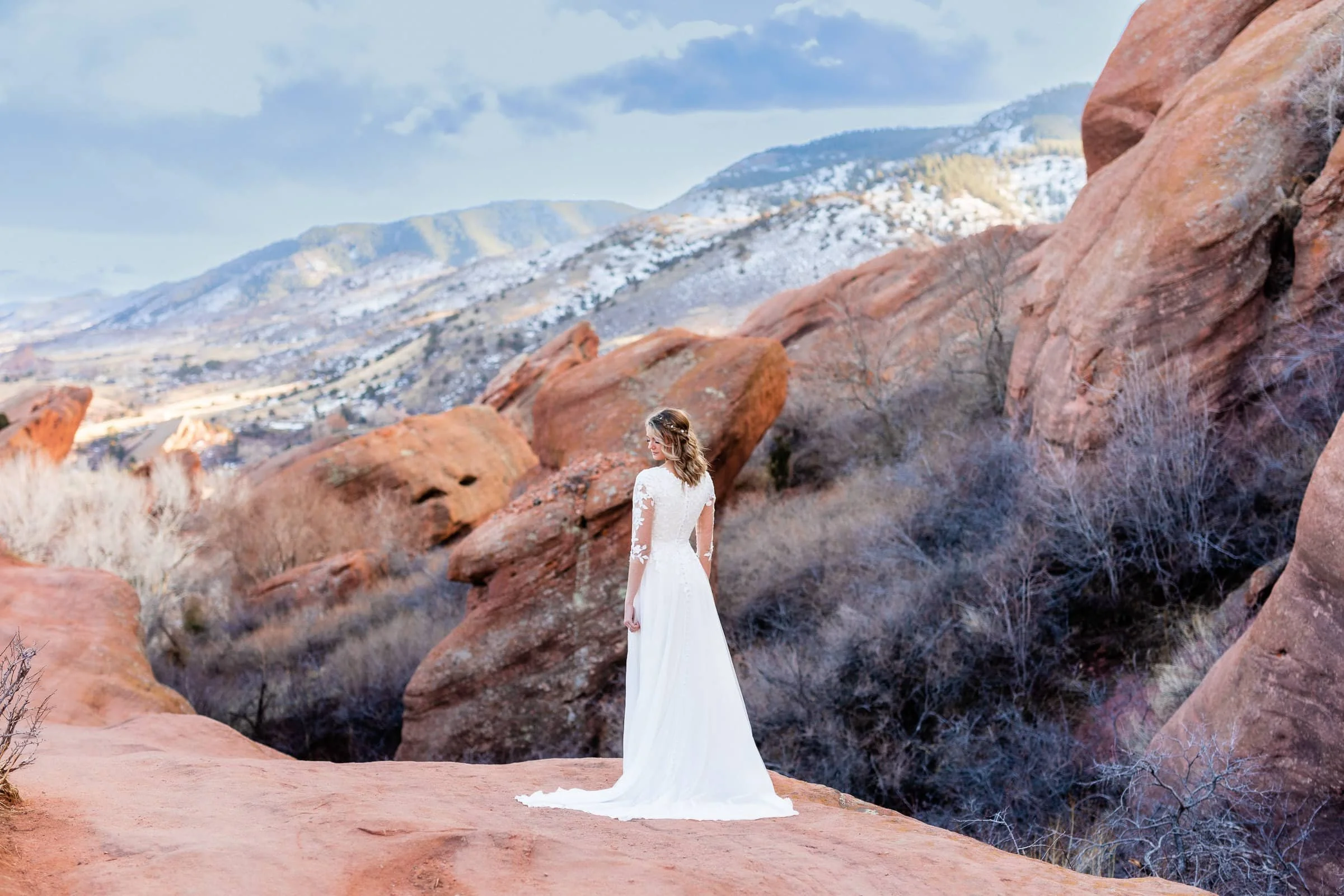 Bride in a white wedding dress standing on a rocky formation in a desert landscape with red rocks and a mountainous background.