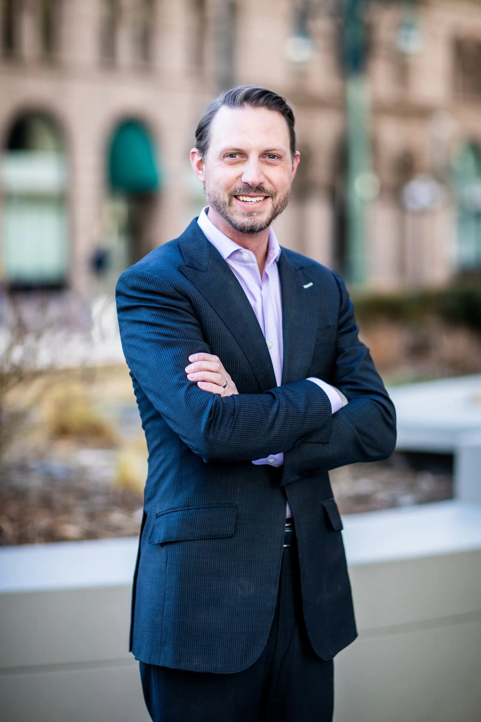 A man in a dark suit, light purple shirt, standing outdoors with arms crossed, smiling, with a blurred background of a building and plants.