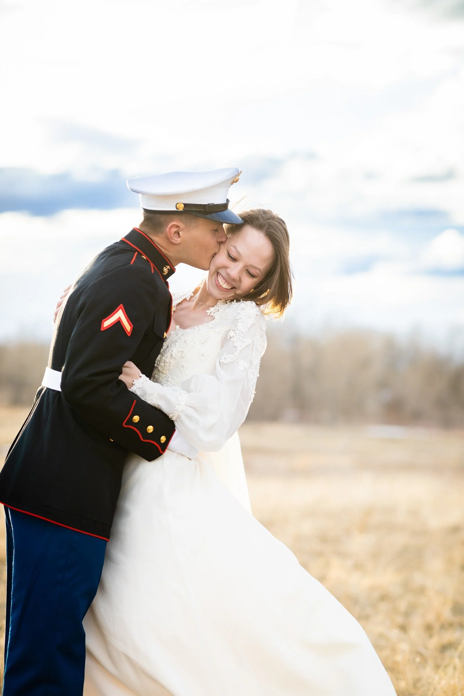 A military man in uniform kissing a smiling woman in a white wedding dress outdoors, with a cloudy sky and field in the background.