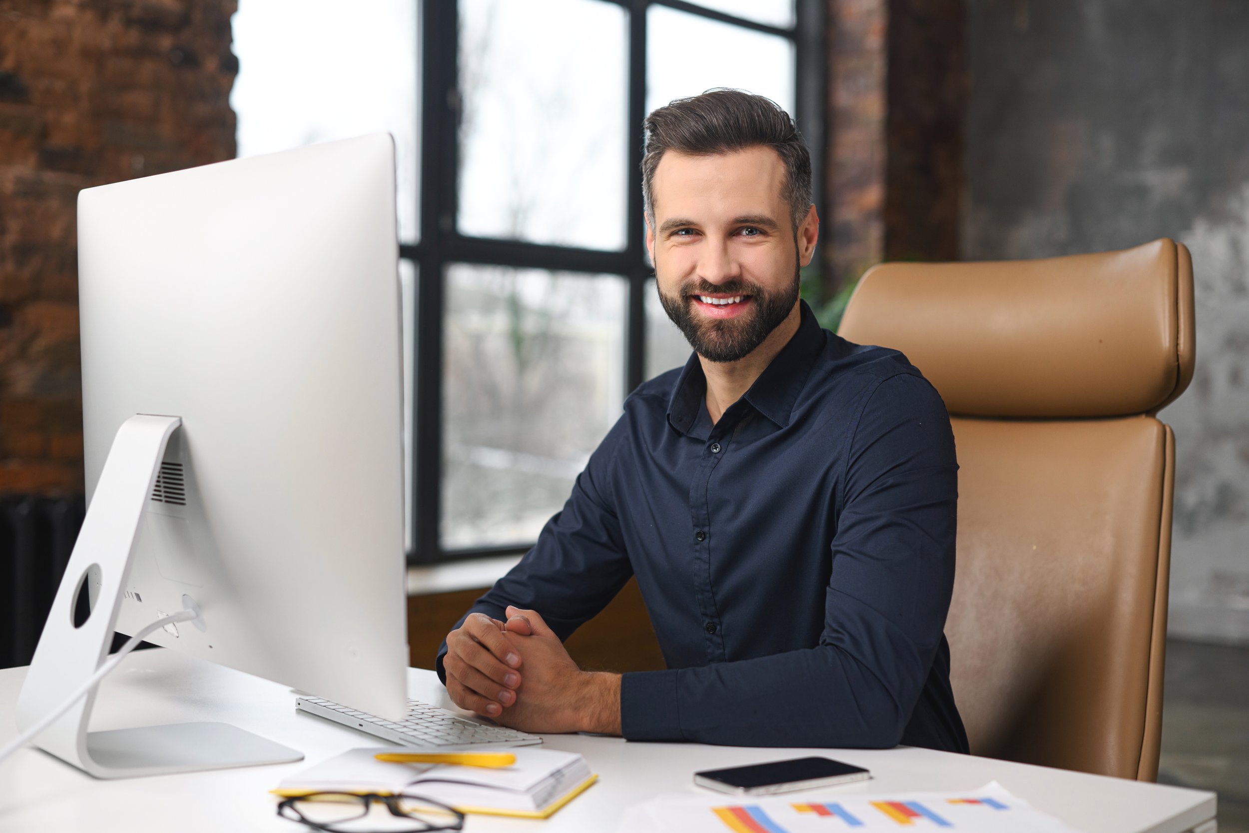 A man with dark hair and a beard, wearing a dark blue shirt, sitting at a white desk in front of a computer monitor in a modern office space with brick walls and large windows, smiling at the camera.