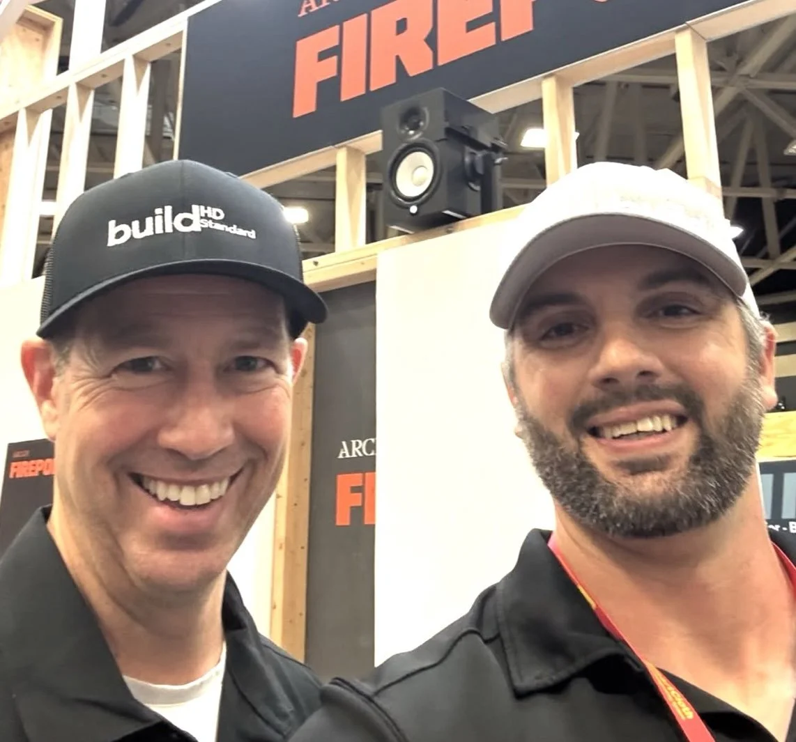 Two smiling men wearing baseball caps and black shirts taking a selfie at a home building trade show, with a large sign that says 'Firepoint' in the background.