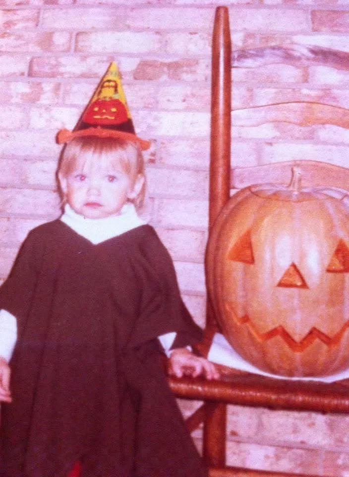 Photo of a young child with dressed as a witch standing next to a wicker chair. In the chair sits a jack-o-lantern.