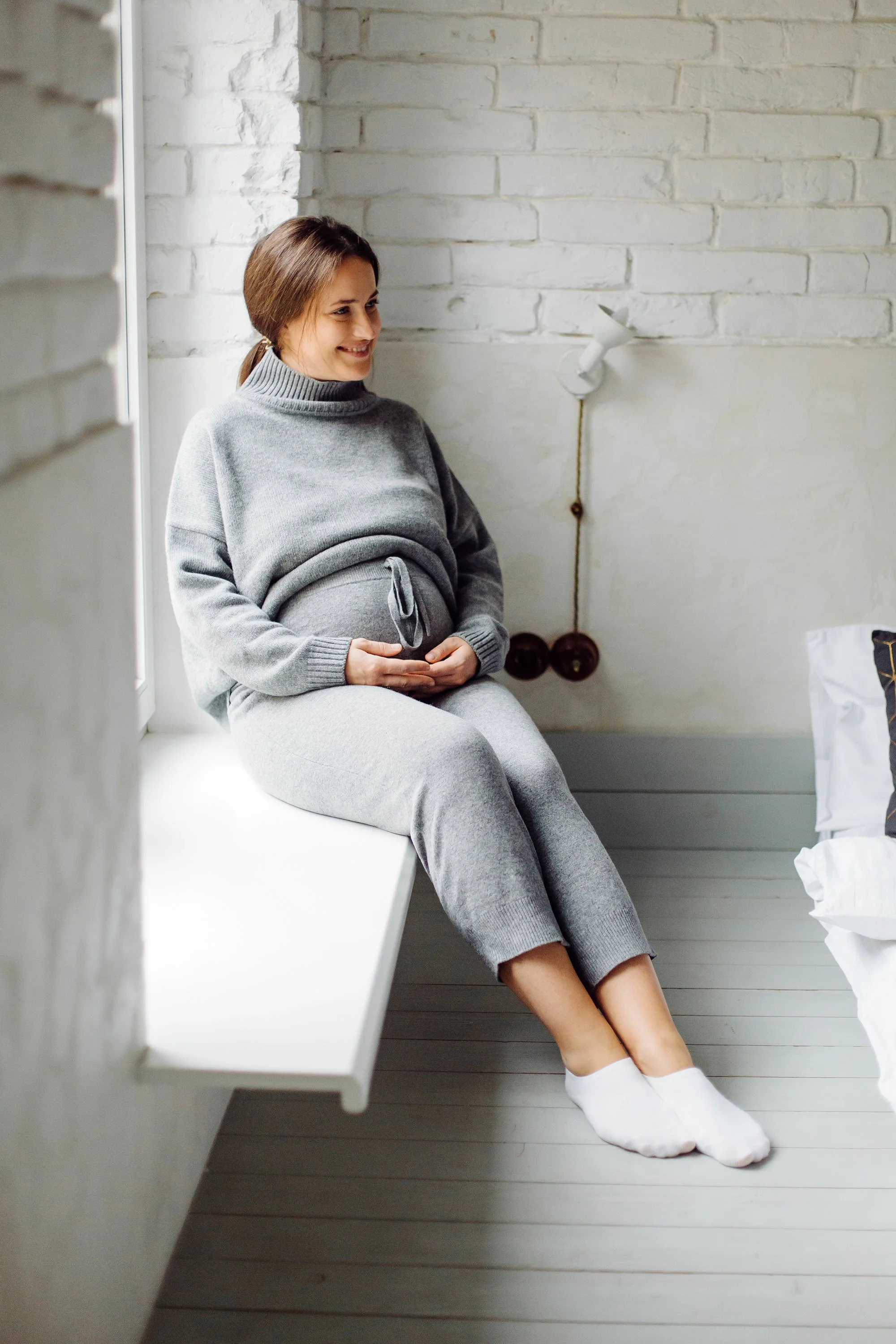 A pregnant woman sitting on a windowsill, smiling, in a gray lounge set, with her hands resting on her belly, in a bright, minimal room.